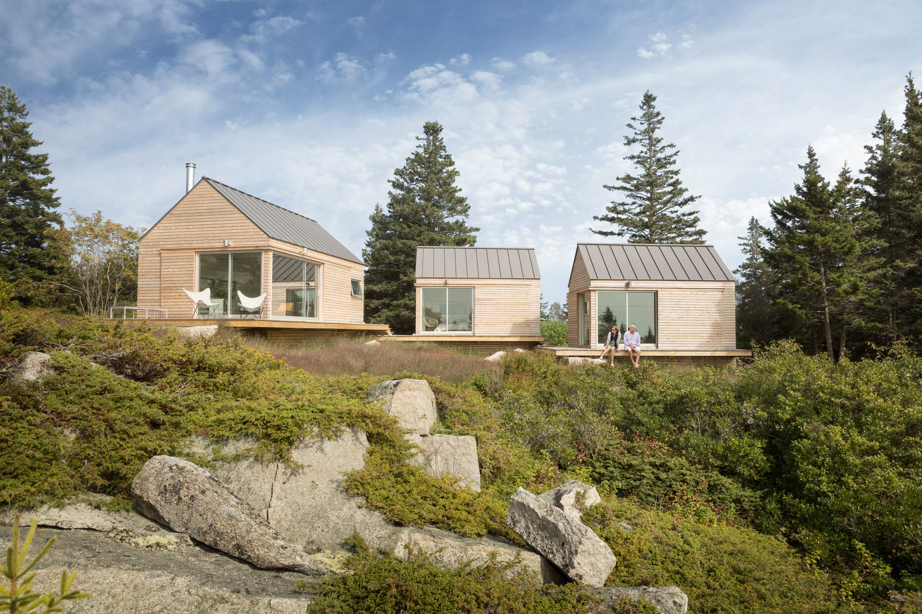 Set on a rocky outcrop and surrounded by evergreens, these three wood-clad cabins feel modern yet rooted in the landscape. Each simple, gabled form is wrapped in natural wood and topped with a metal roof, blending seamlessly with the forest and stone.
Sliding glass doors open to wide views and soft light, while a low wooden platform links the cabins as both walkway and gathering space. The design is quiet and deliberate, letting the rugged setting speak while offering a calm place to stay.