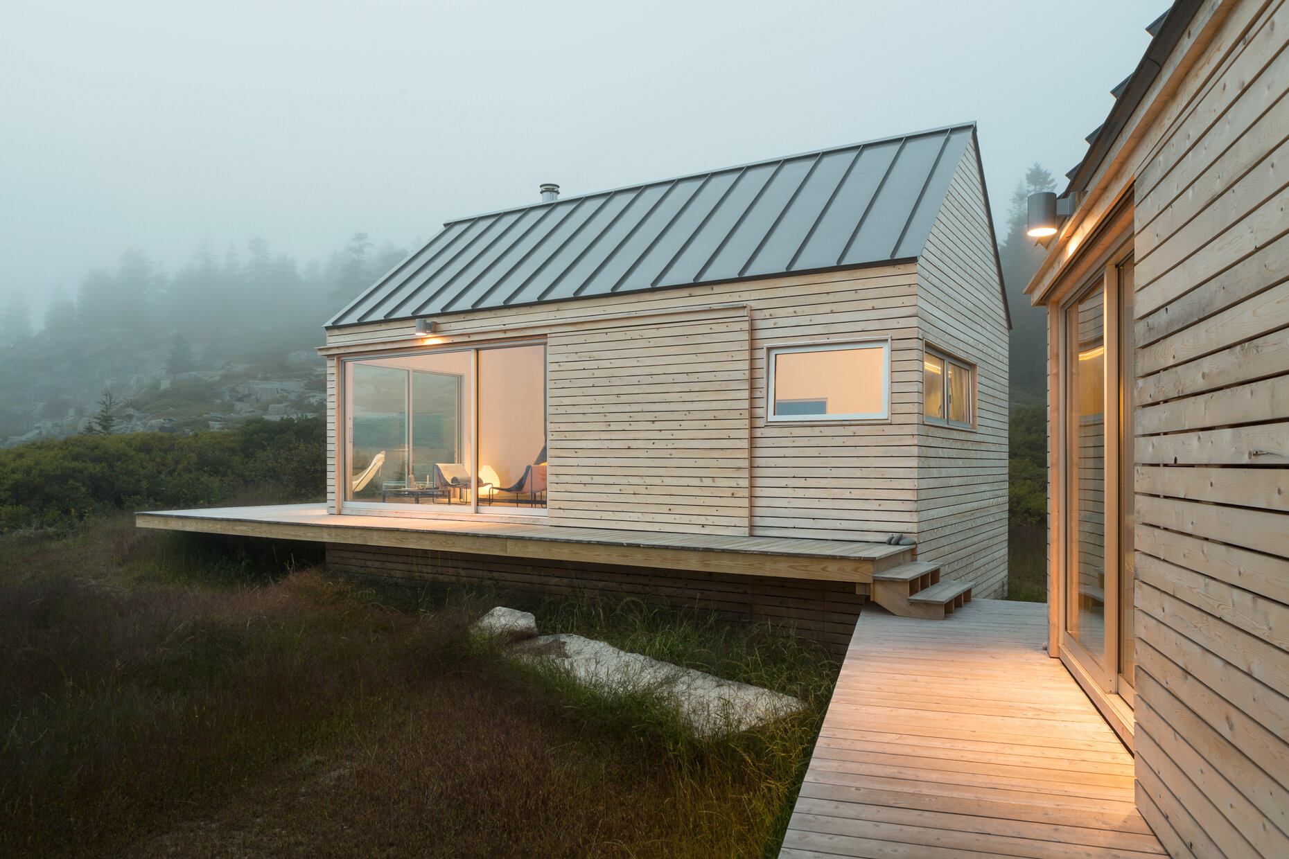 Set on a rocky outcrop and surrounded by evergreens, these three wood-clad cabins feel modern yet rooted in the landscape. Each simple, gabled form is wrapped in natural wood and topped with a metal roof, blending seamlessly with the forest and stone.
Sliding glass doors open to wide views and soft light, while a low wooden platform links the cabins as both walkway and gathering space. The design is quiet and deliberate, letting the rugged setting speak while offering a calm place to stay.