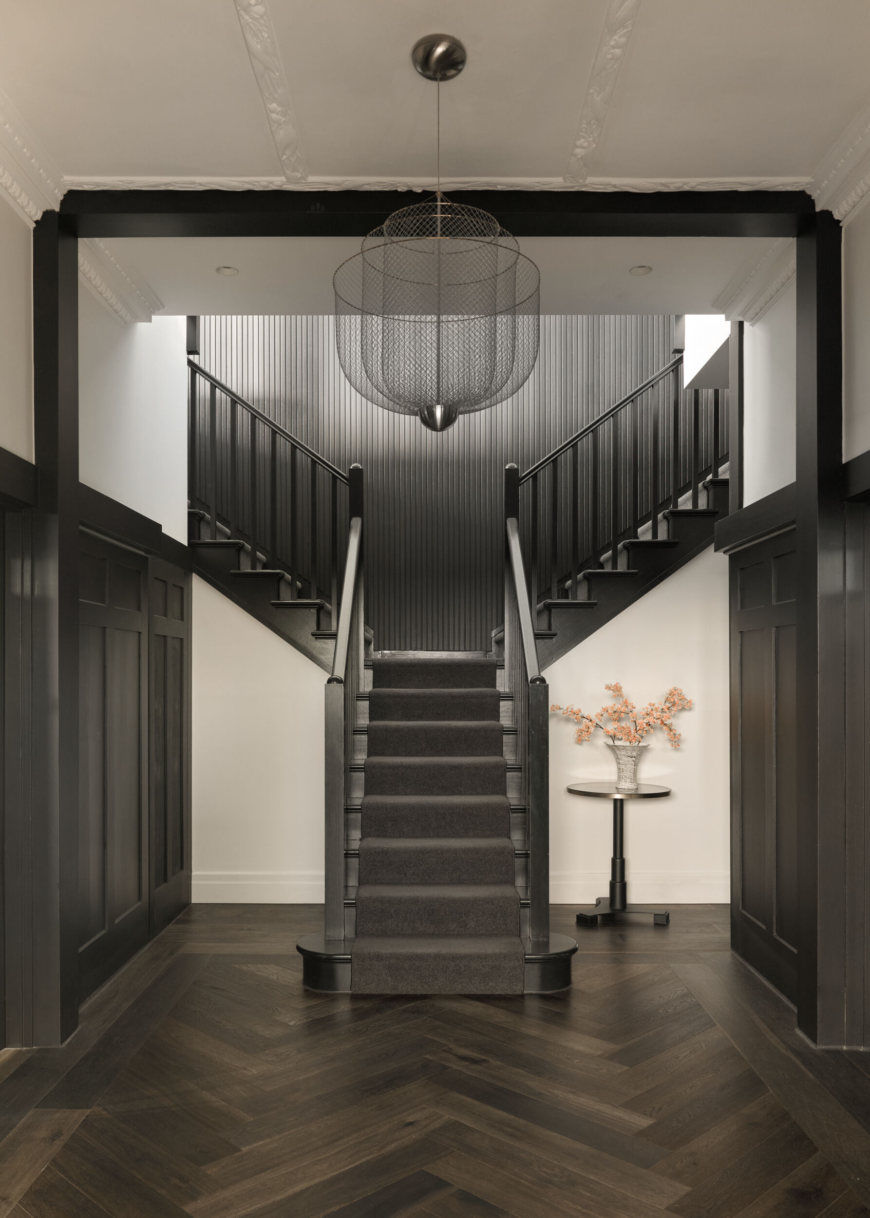 This staircase makes a dramatic first impression, pairing architectural grandeur with sculptural subtlety. The split staircase rises like a stage set, symmetrically framed by sleek black balustrades and a vertical black accent wall that draws the eye upward. The floor, laid in a rich herringbone pattern, adds movement and warmth underfoot while echoing the home’s Deco roots with a refined sense of rhythm.