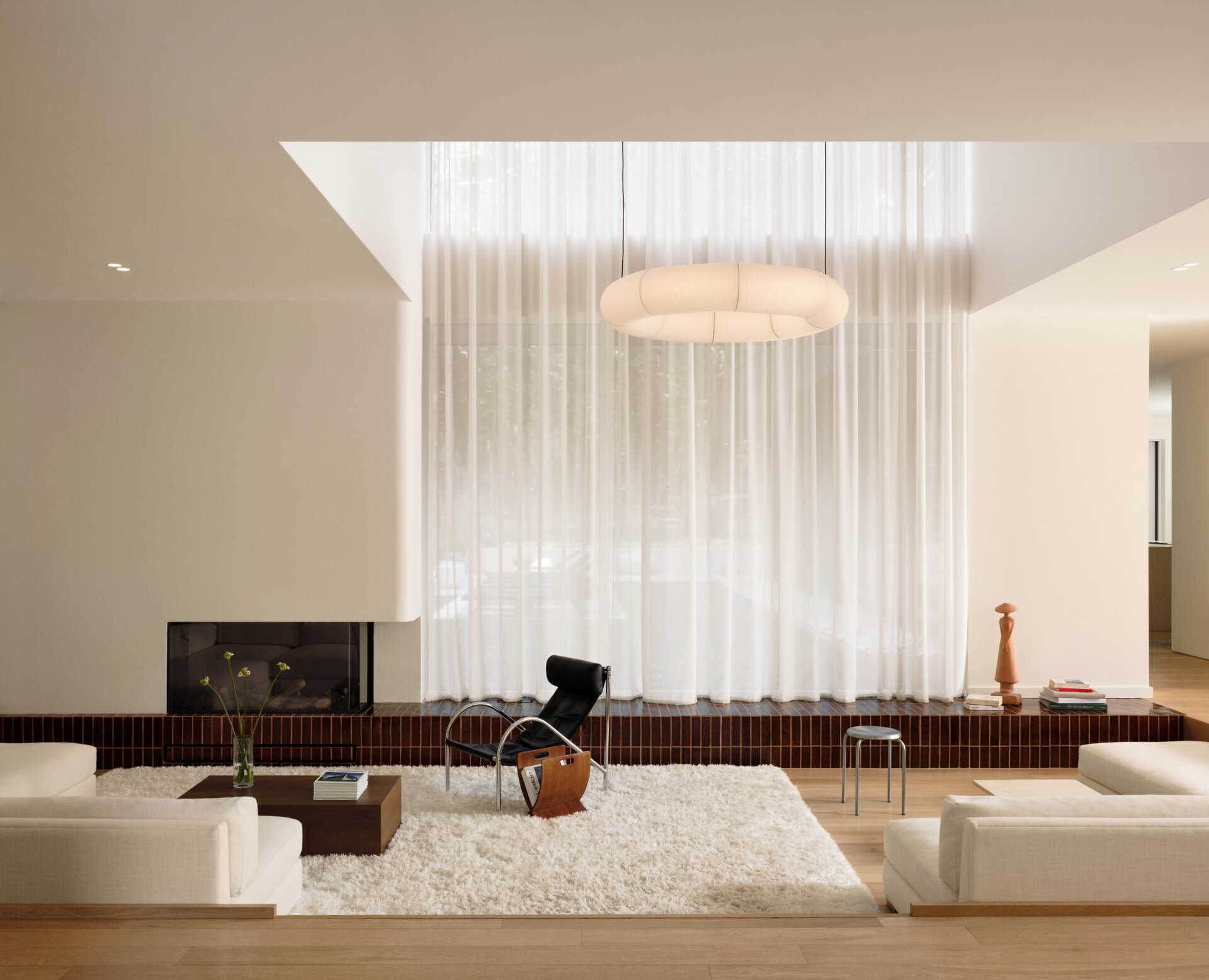 This sunken living room is both sculptural and serene. It creates an inviting, grounded space centered around a plush ivory rug and a terracotta tile platform. Rich in tone and laid with clean precision, the tile provides a grounding base for both the fireplace and the window wall.  Soft curves in the white plaster echo the home's rounded details, while pale sofas and a black leather lounge chair add contrast and calm. A sheer curtain highlights the double-height ceiling, and a glowing washi paper pendant adds warmth.