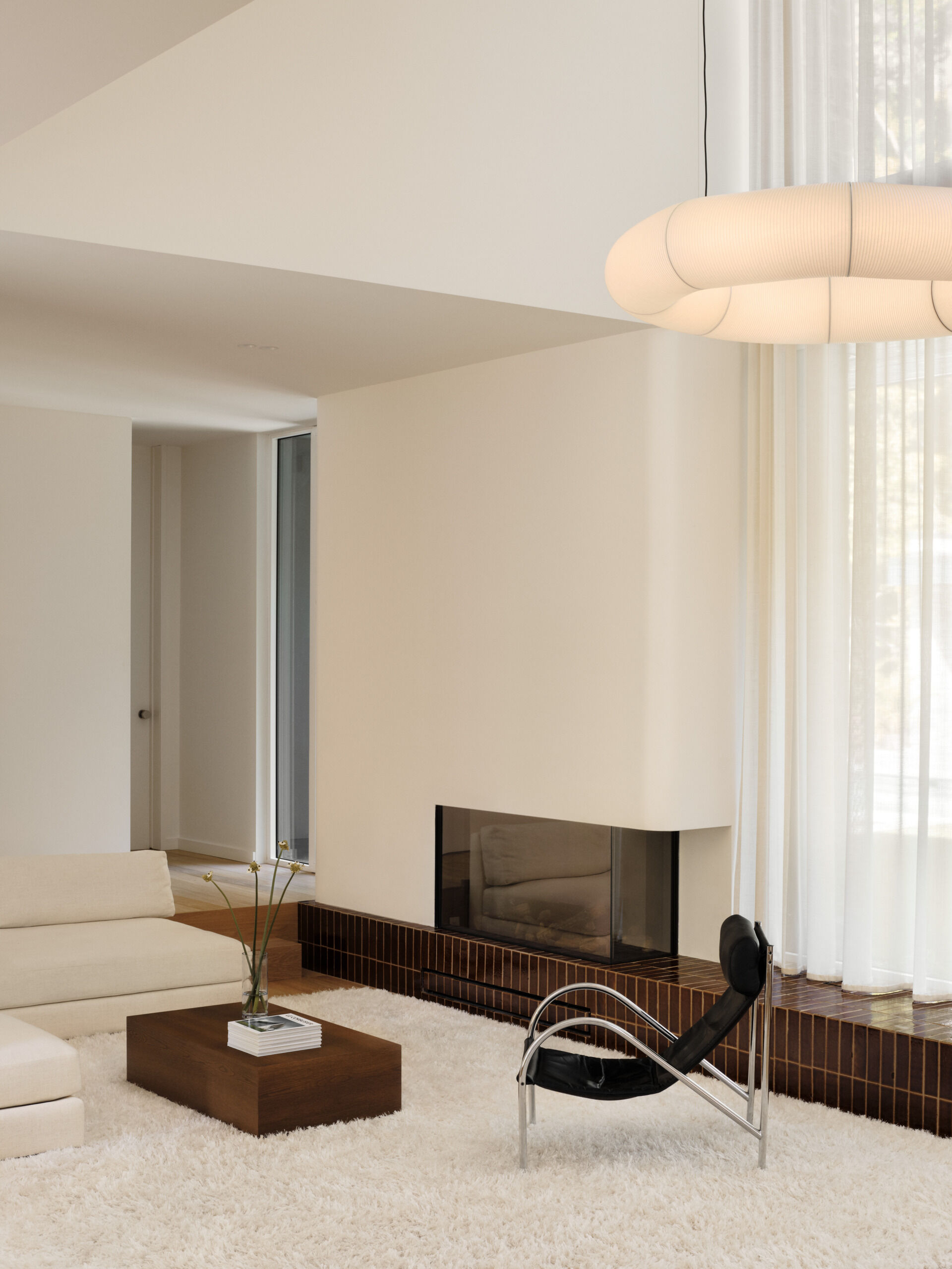 This sunken living room is both sculptural and serene. It creates an inviting, grounded space centered around a plush ivory rug and a terracotta tile platform. Rich in tone and laid with clean precision, the tile provides a grounding base for both the fireplace and the window wall.  Soft curves in the white plaster echo the home's rounded details, while pale sofas and a black leather lounge chair add contrast and calm. A sheer curtain highlights the double-height ceiling, and a glowing washi paper pendant adds warmth.