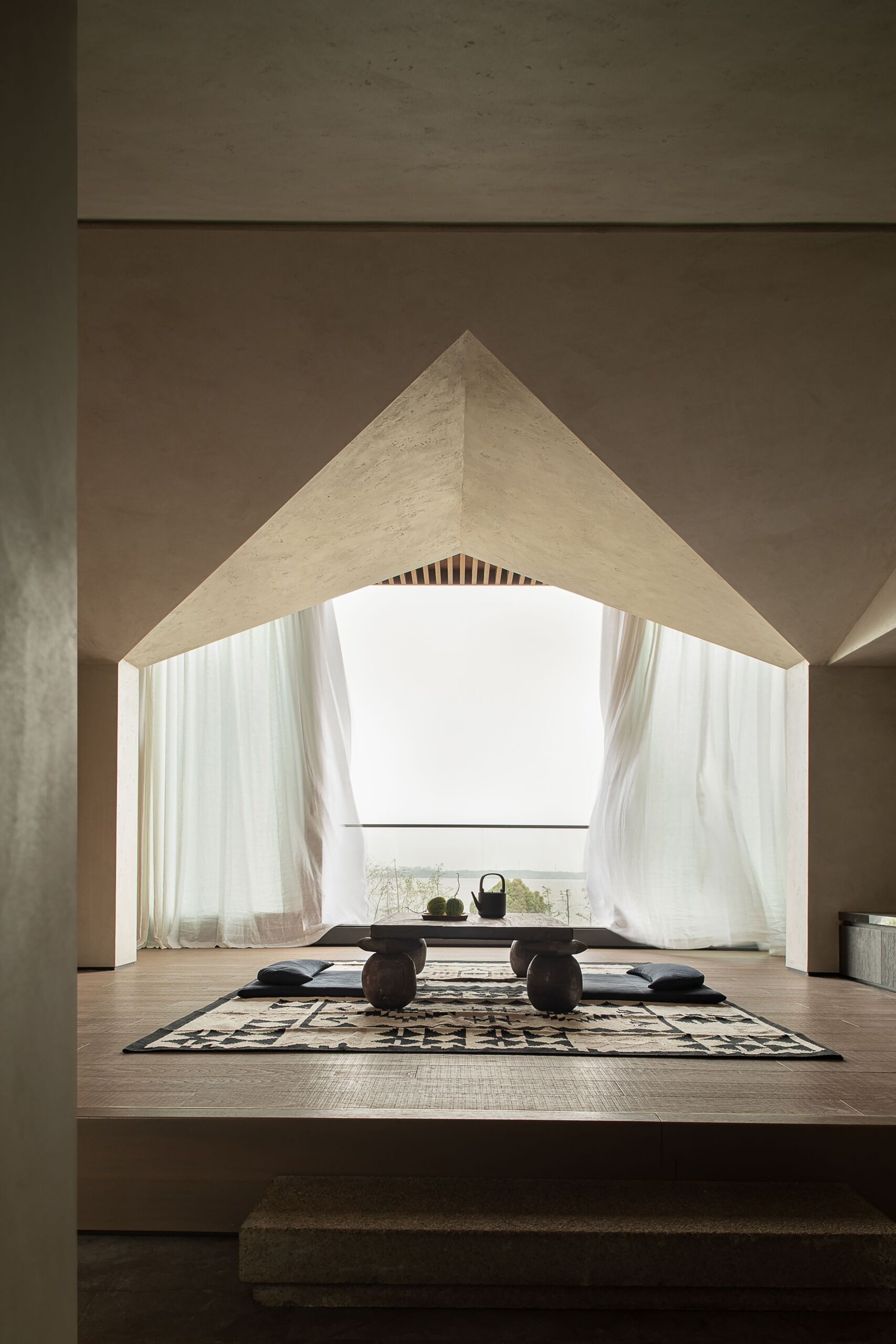 Bathed in soft daylight, this elevated tea room feels like a quiet refuge suspended in the sky. A low wooden table rests on a textured woven rug, creating a cozy focal point grounded in natural tones. Billowing white curtains frame the space with a sense of movement, while the sharply sculpted ceiling draws the eye upward. Every element invites stillness and clarity, making this a perfect setting for mindful moments and slow sips with a view.