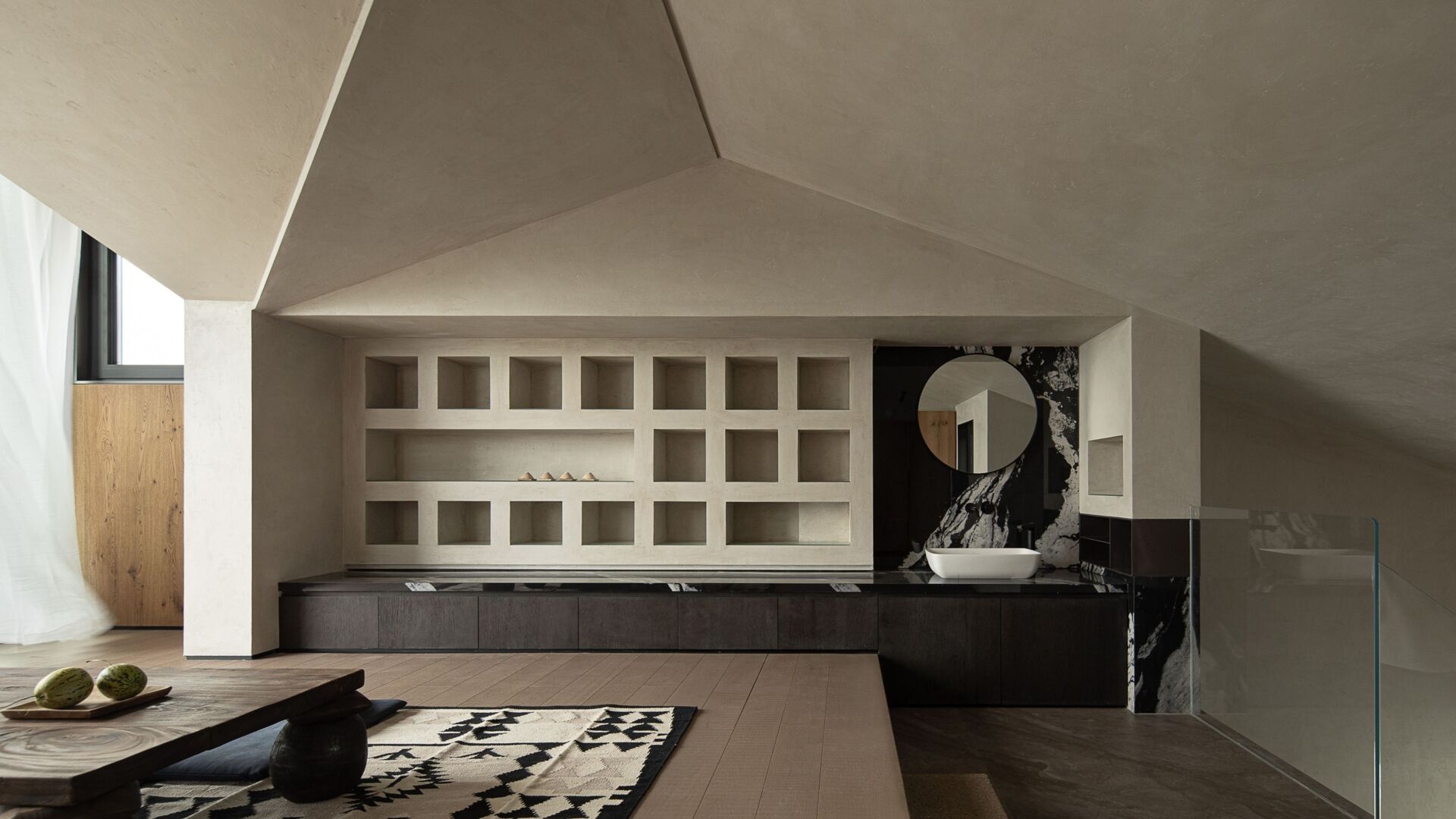 Clean geometry and natural materials come together in this serene alcove, where a grid of built-in shelves adds quiet rhythm to the space. Just beyond the tea platform, a black-and-white marble backsplash anchors the small area, its round mirror echoing the room’s soft, sculptural forms. The ceiling folds overhead like origami, casting gentle shadows that shift with the light.