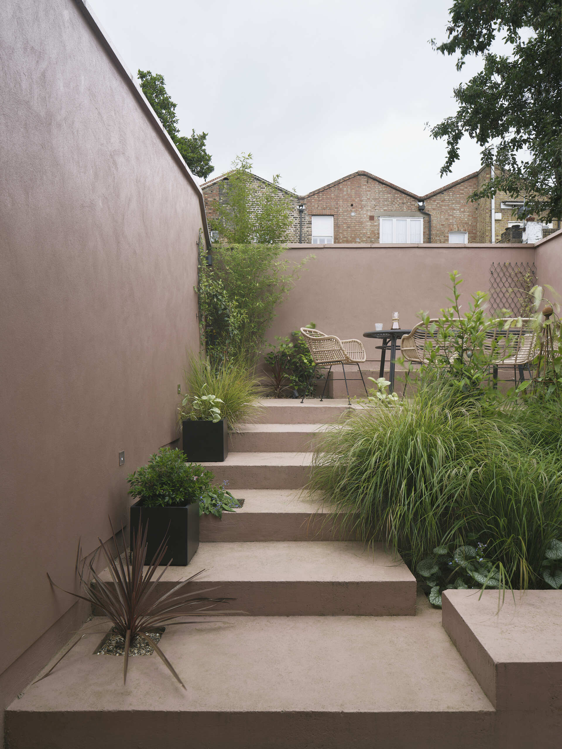 This terrace garden becomes the focal point of the home, with a series of steps and built-in planters guiding users to the top, where a private elevated space features a table and chairs, offering a quiet spot to relax or entertain, surrounded by walls and greenery. This layered outdoor area continues the home’s theme of connected, purposeful spaces.