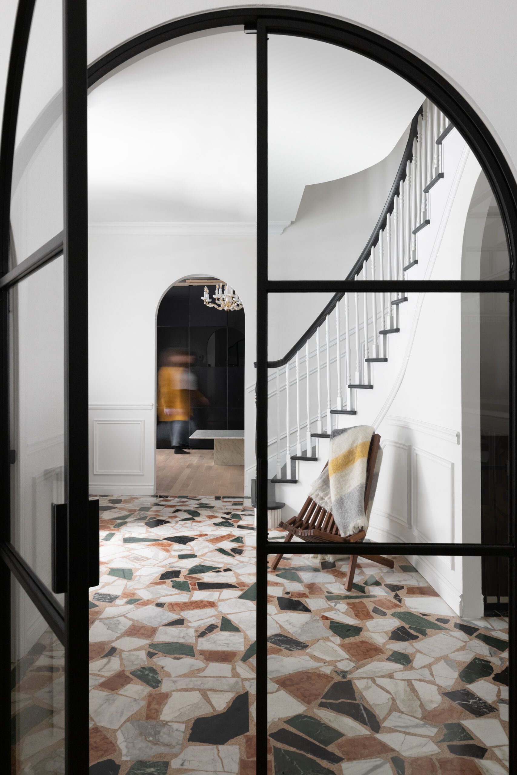 A glass-paned steel door opens into a crisp white foyer, where a curved staircase with slender white spindles and a dark wooden handrail anchors the space. A low wooden chair sits quietly at the base of the stairs, softened by a striped throw. Underfoot, large terrazzo-style tiles in earthy tones, terracotta, green, cream, and black, create a striking patchwork floor that feels both contemporary and timeless.