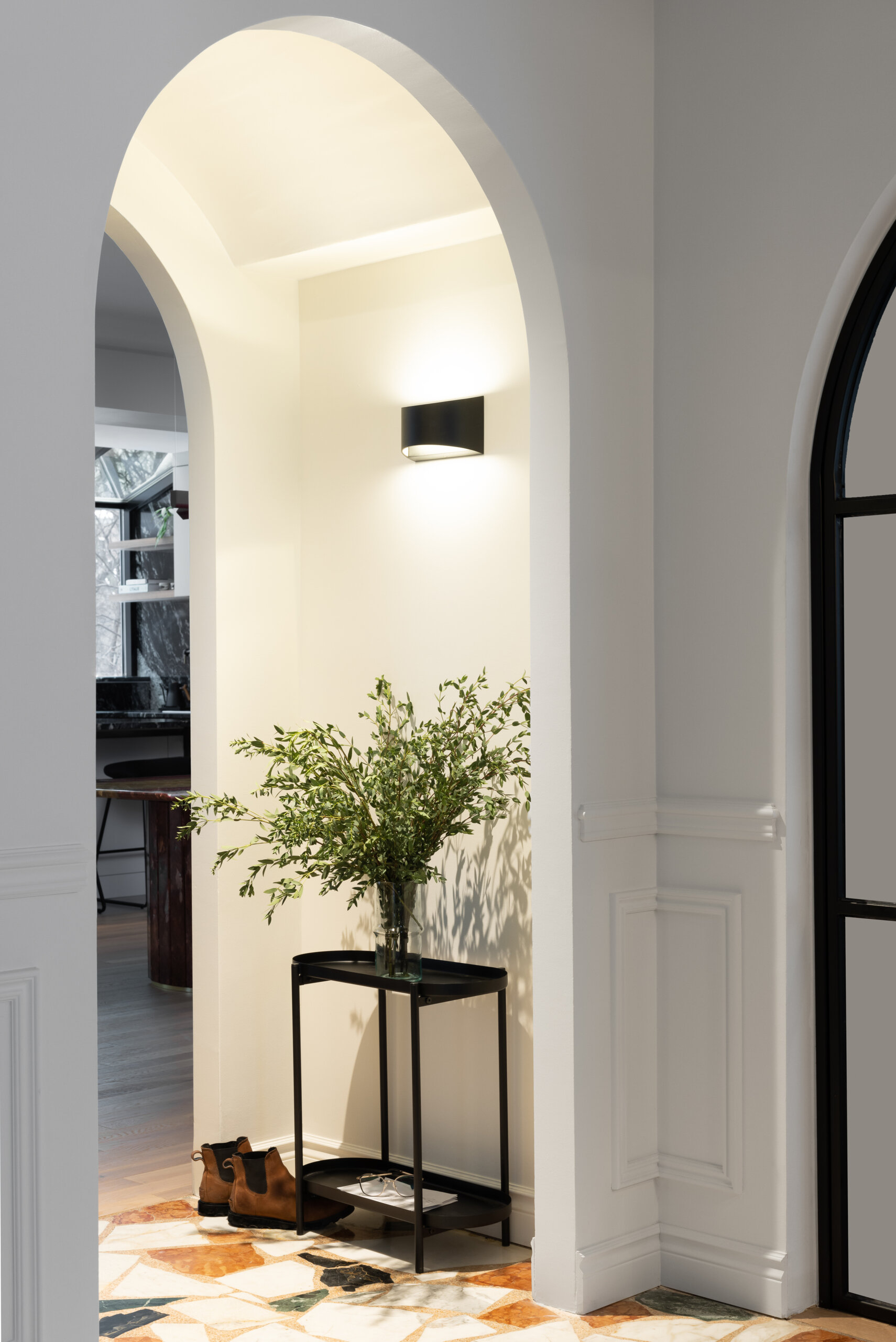 A glass-paned steel door opens into a crisp white foyer, where a curved staircase with slender white spindles and a dark wooden handrail anchors the space. A low wooden chair sits quietly at the base of the stairs, softened by a striped throw. Underfoot, large terrazzo-style tiles in earthy tones, terracotta, green, cream, and black, create a striking patchwork floor that feels both contemporary and timeless.