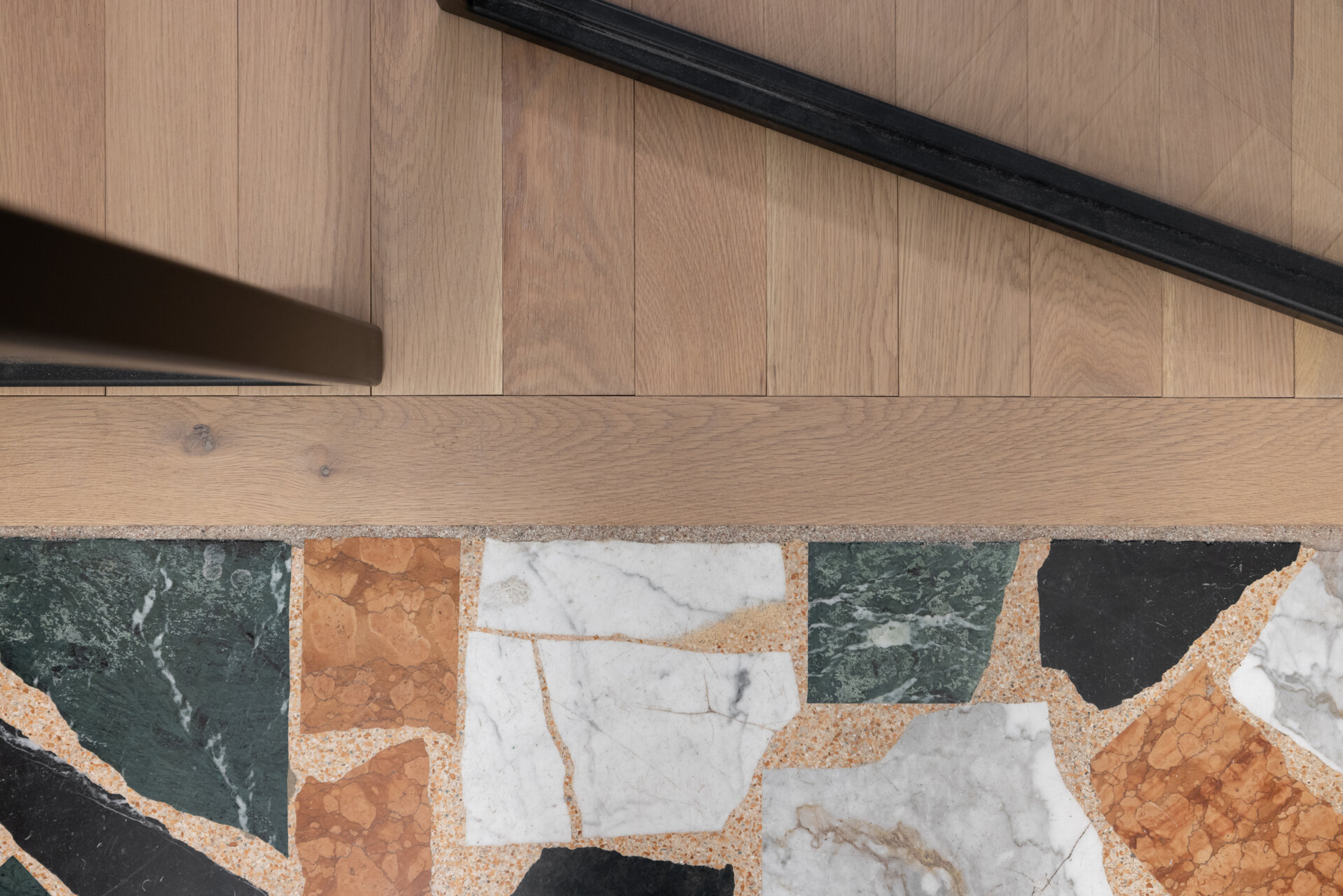 A glass-paned steel door opens into a crisp white foyer, where a curved staircase with slender white spindles and a dark wooden handrail anchors the space. A low wooden chair sits quietly at the base of the stairs, softened by a striped throw. Underfoot, large terrazzo-style tiles in earthy tones, terracotta, green, cream, and black, create a striking patchwork floor that feels both contemporary and timeless.
