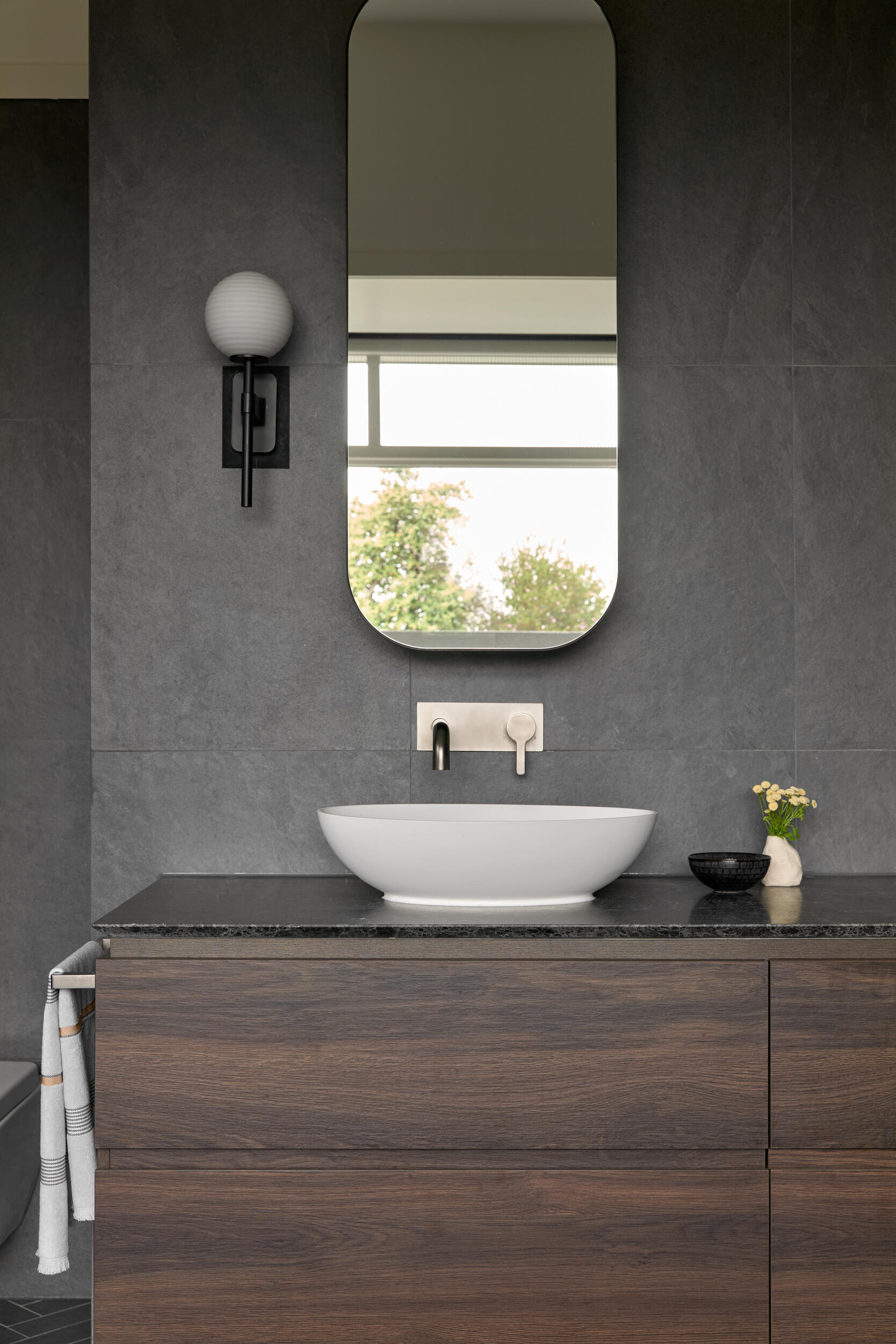 In a bathroom, the vanity features a warm wood base with a simple, matte finish, paired with a crisp white basin that has a rounded, modern shape. The vanity sits in front of dark gray oversized wall tiles, which provide contrast and help anchor the space. Above, a pill-shaped mirror adds softness to the overall geometry, while a globe sconce offers functional lighting with a subtle retro touch.