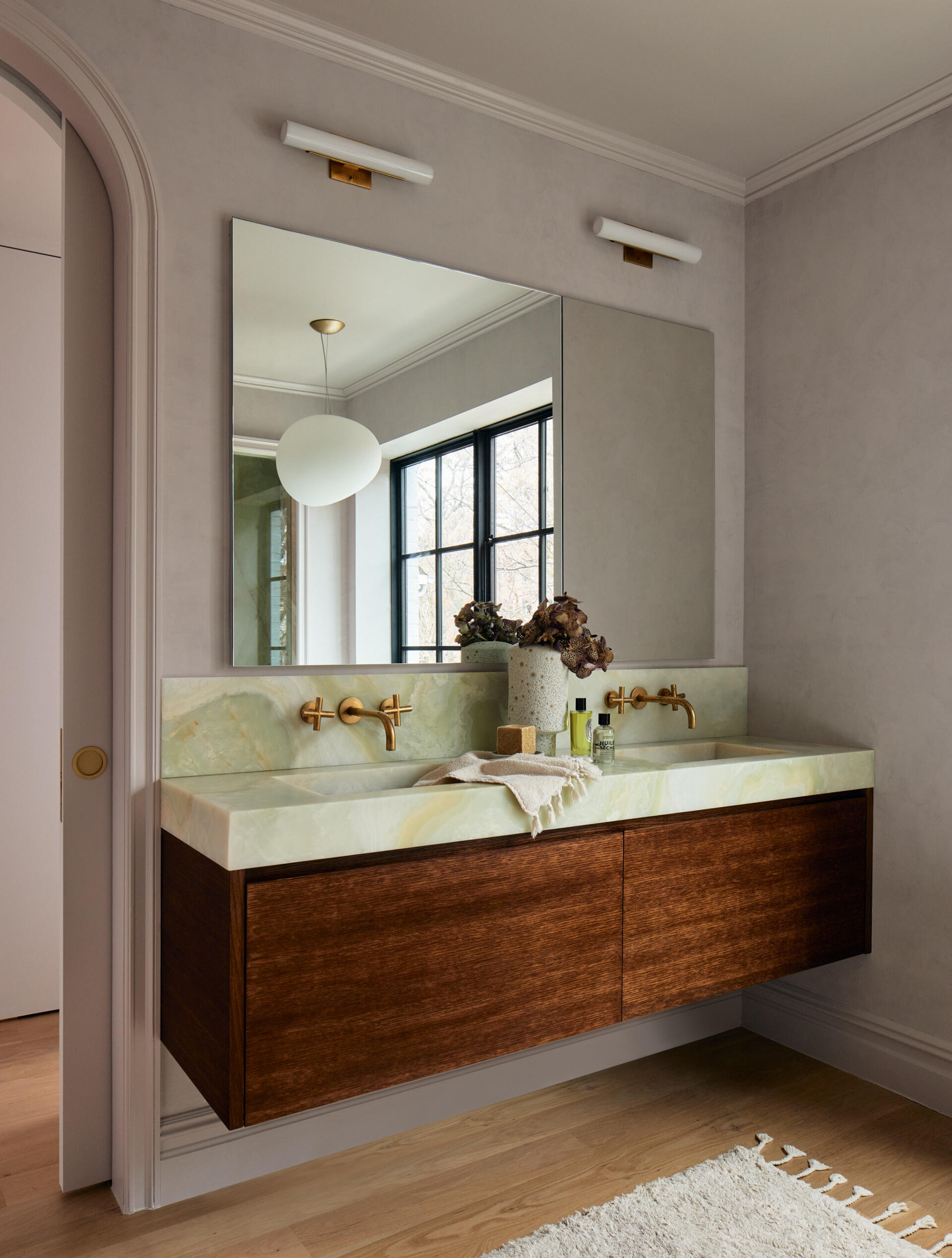 This primary bathroom continues the lavender-gray plaster from the bedroom and elevates it with dramatic stone selections. Green onyx and rosa aurora macchia vecchia marble define the shower, while a freestanding tub sits beneath a steel casement window overlooking the garden and the wood vanity is topped with onyx.
