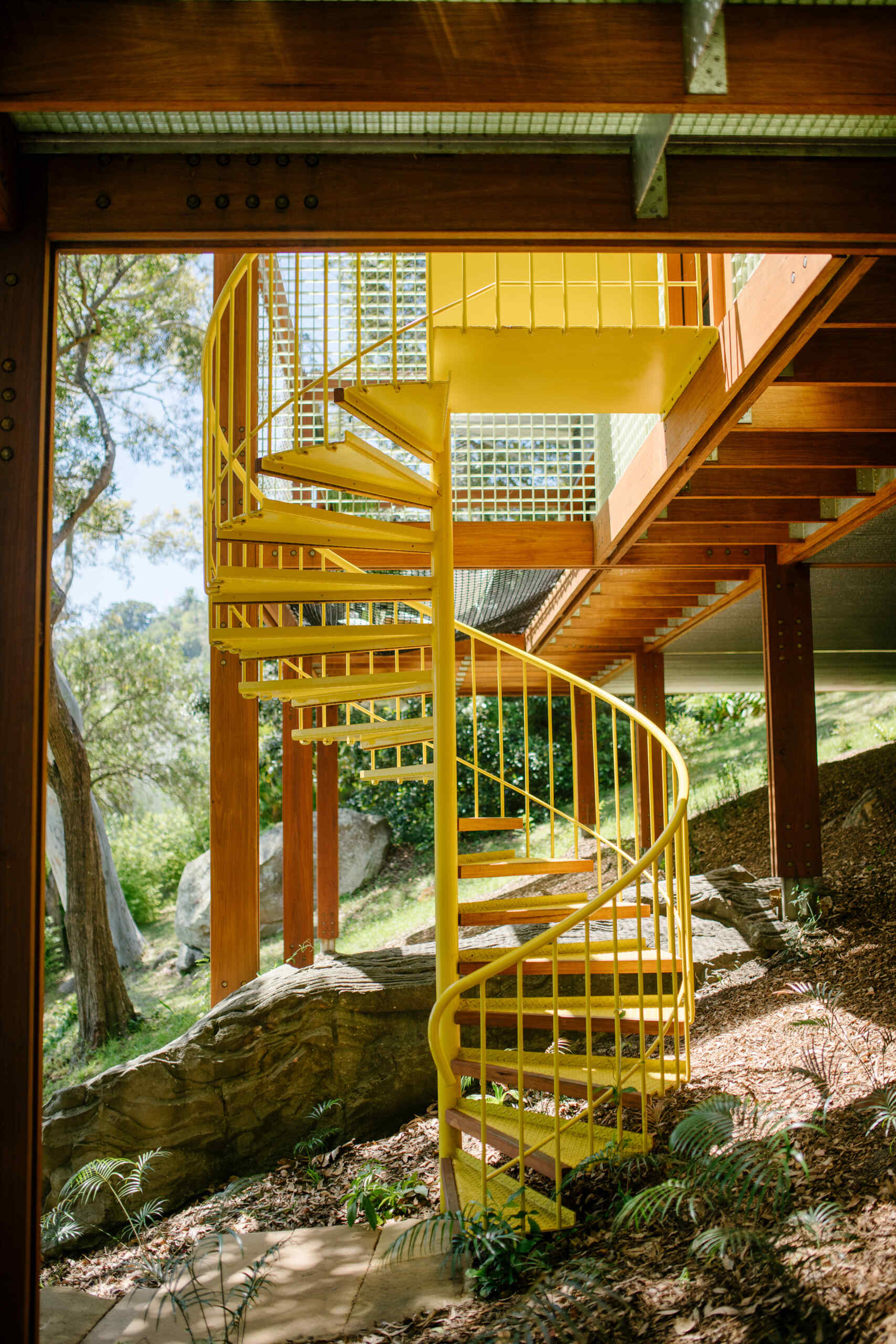 A bold yellow spiral staircase winds up through the stone and foliage, adding a playful sculptural twist to this quiet treetop home.