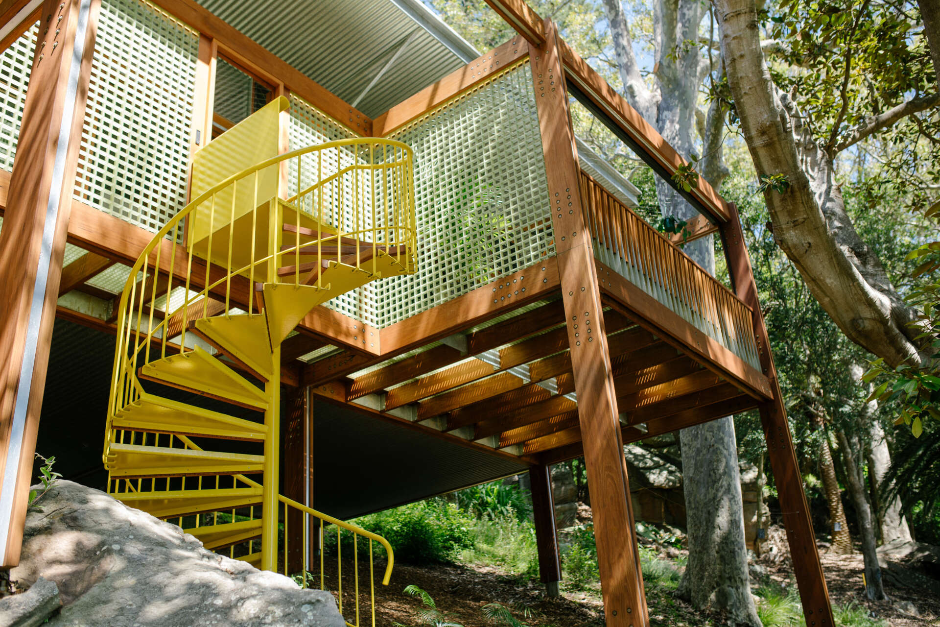A bold yellow spiral staircase winds up through the stone and foliage, adding a playful sculptural twist to this quiet treetop home.