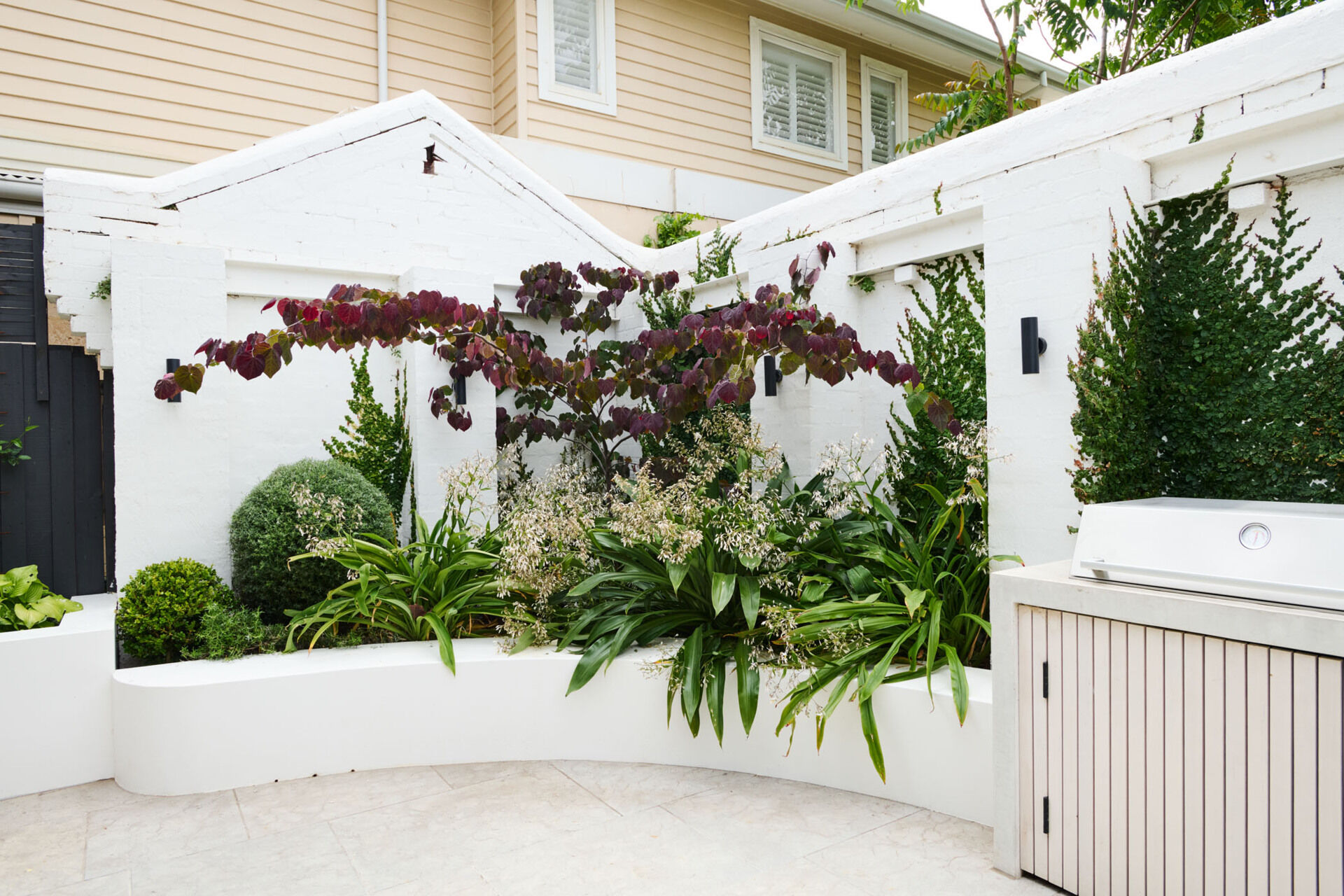 This tranquil alfresco area transforms a former garage wall into a cozy garden retreat. Layered planting, a timber pergola, and a built-in BBQ make it perfect for relaxing or entertaining outdoors.