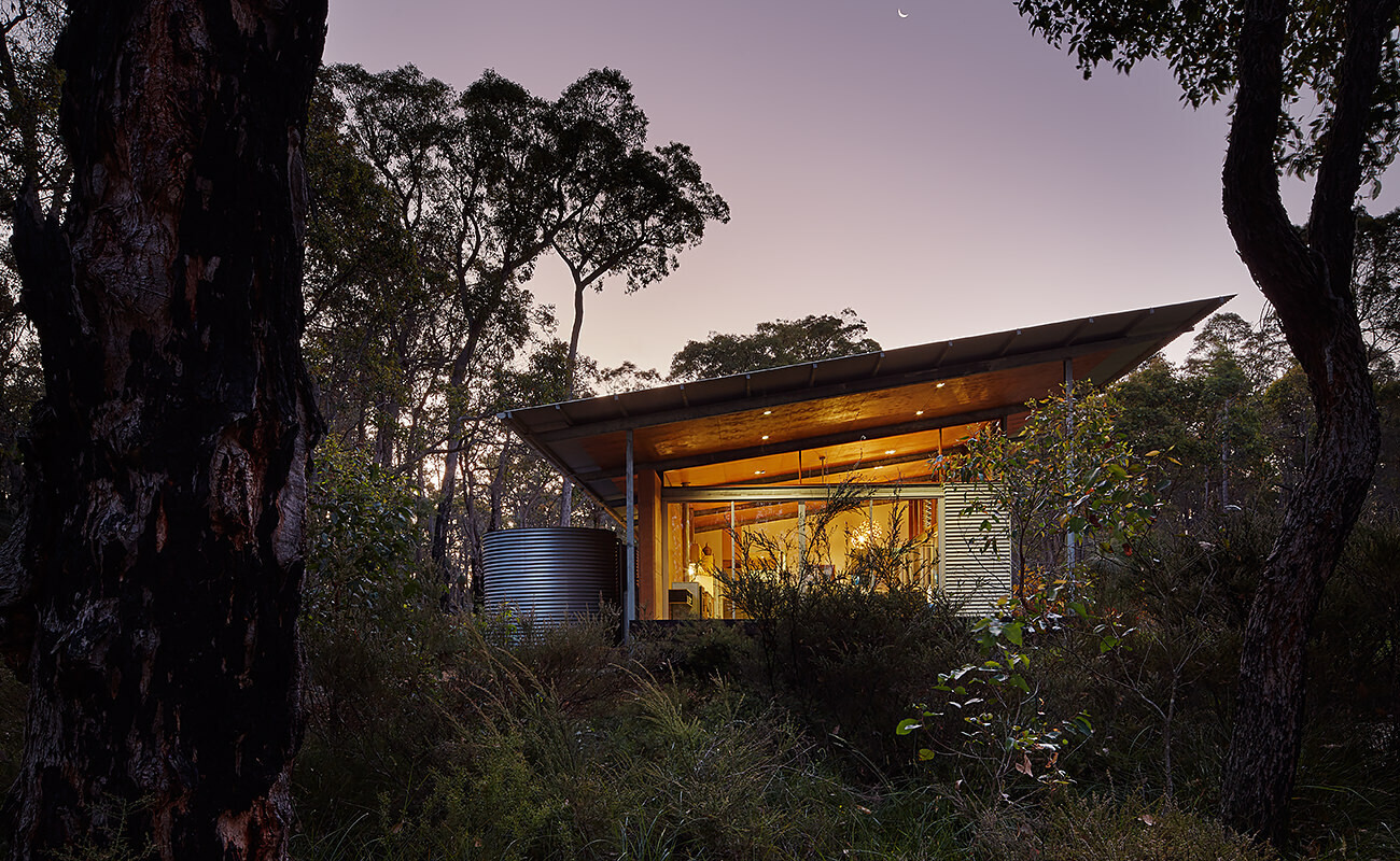 A bushland pavilion with exposed steel frames, rammed earth walls, and a single sweeping roof, inspired by mid-century modern design.