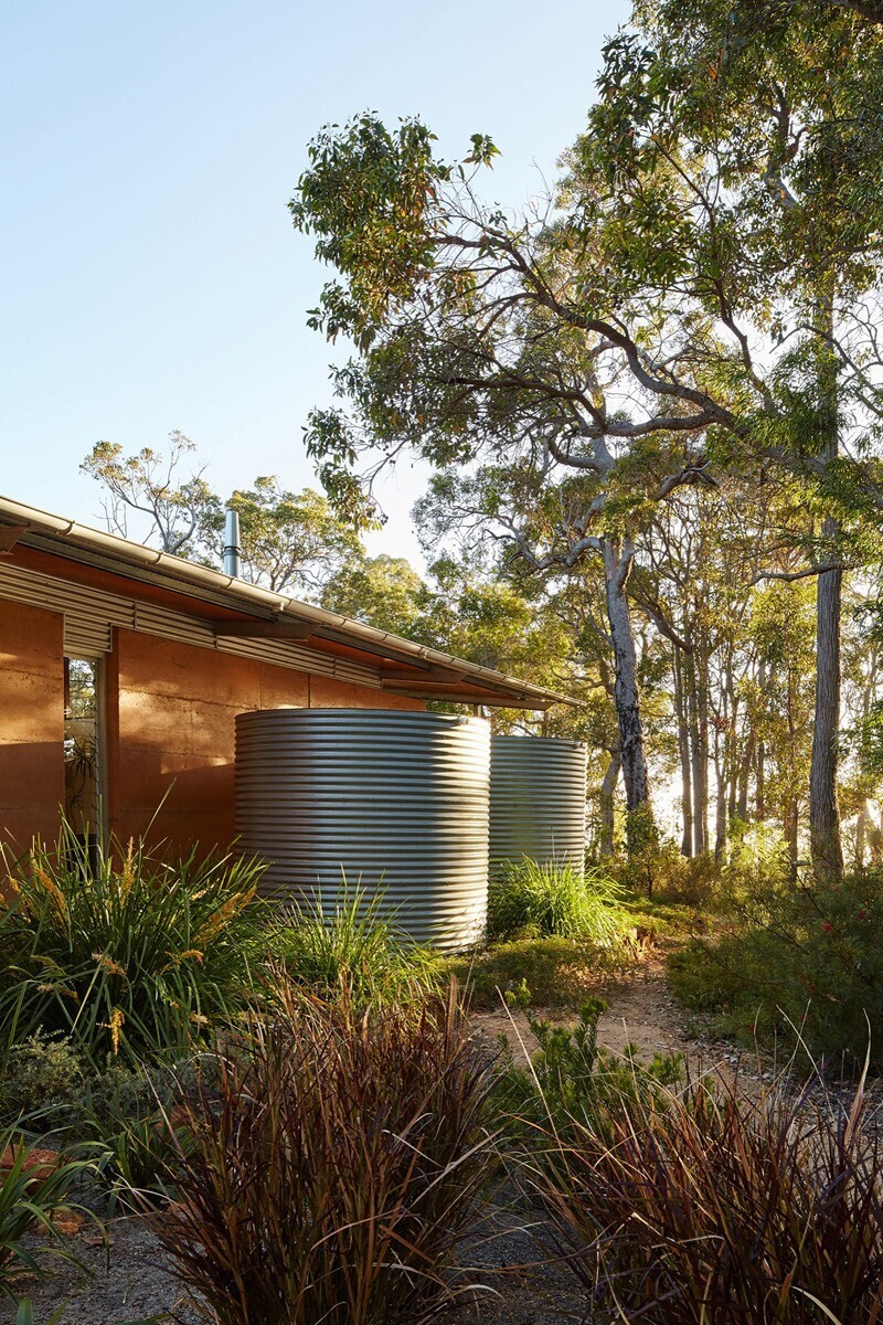 A home in the bush with exposed steel frames, rammed earth walls, and a single sweeping roof, inspired by mid-century modern design.