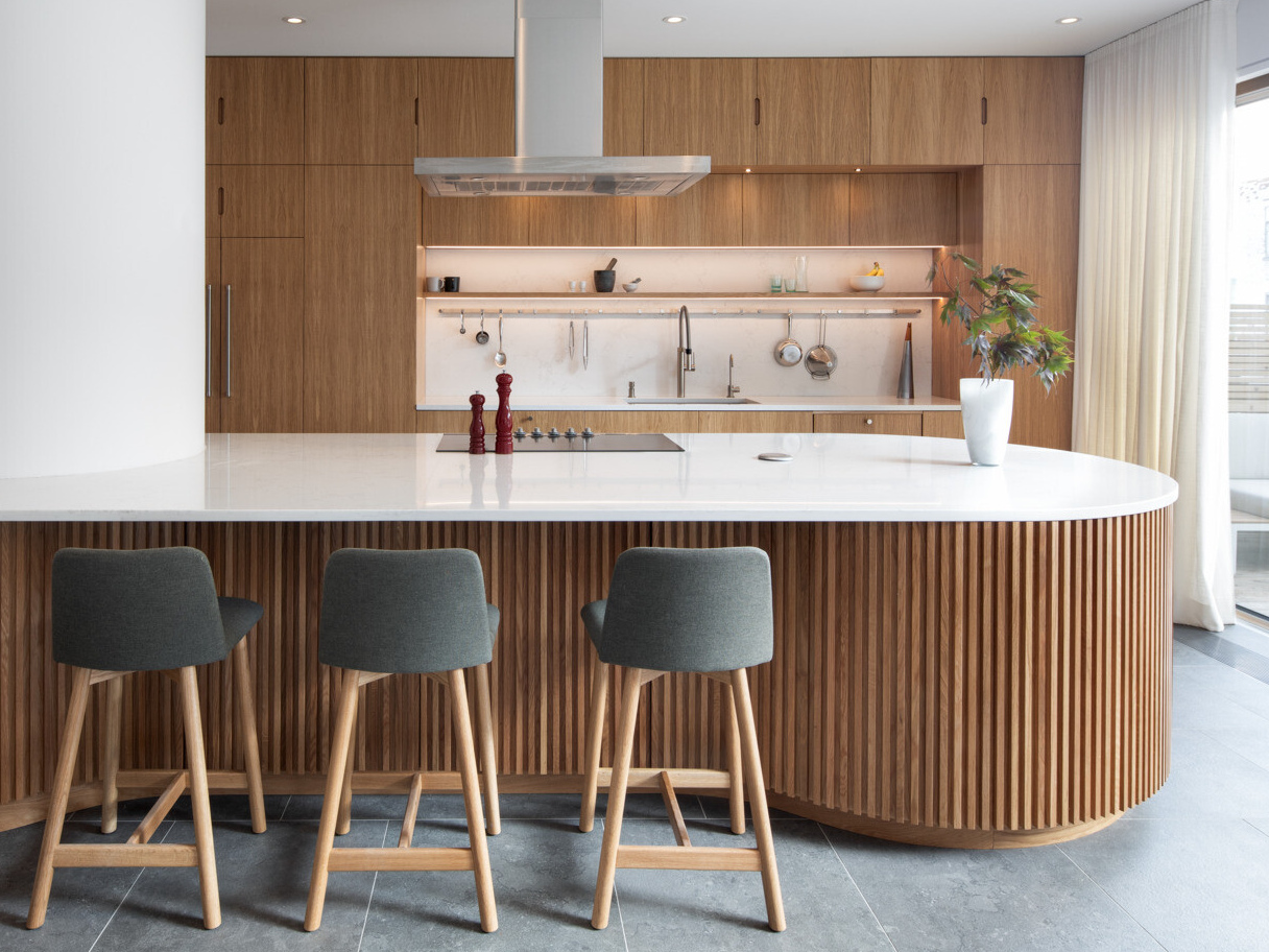 A sculptural oak island and seamless floor-to-ceiling cabinetry define this light-filled Montreal kitchen, where natural materials and soft curves create a warm, minimalist atmosphere.