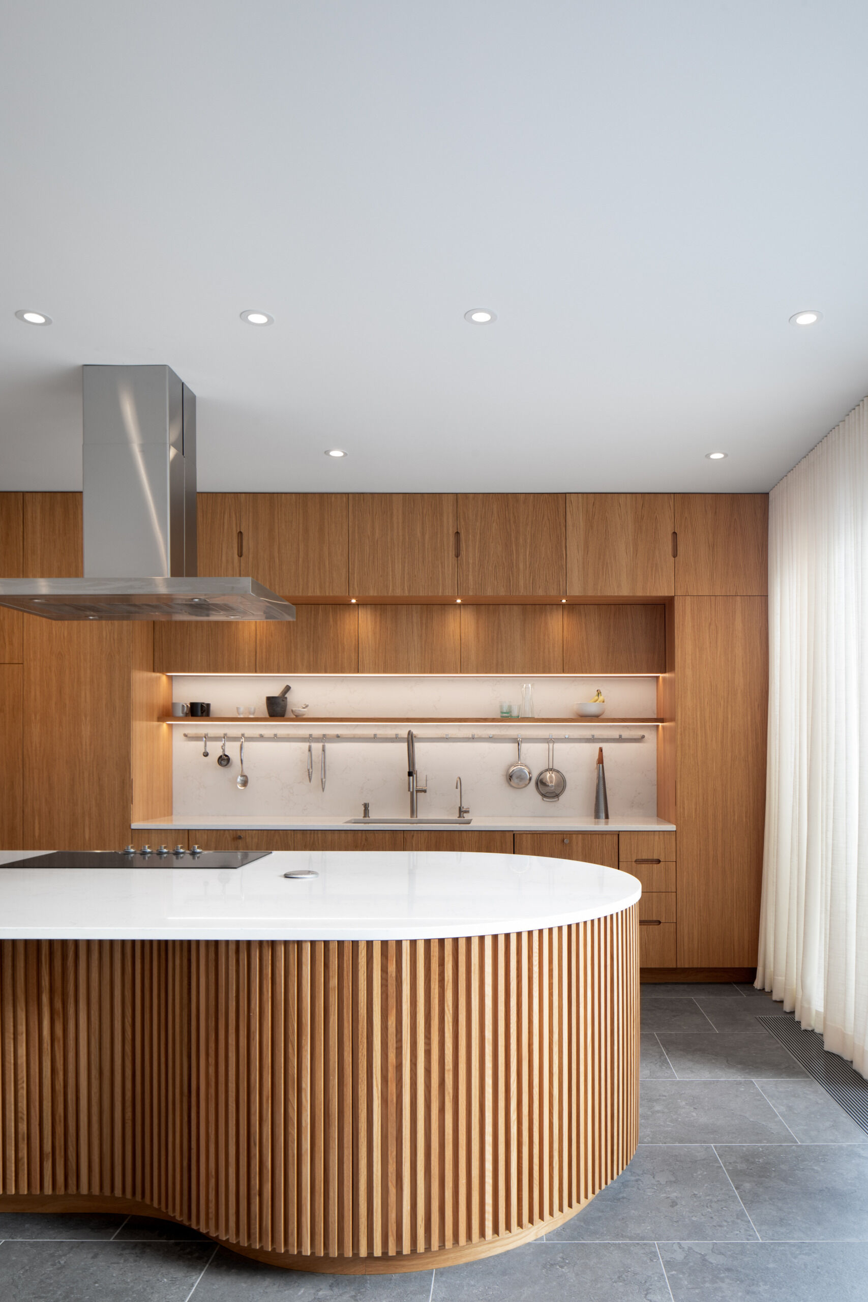 A sculptural oak island and seamless floor-to-ceiling cabinetry define this light-filled Montreal kitchen, where natural materials and soft curves create a warm, minimalist atmosphere.