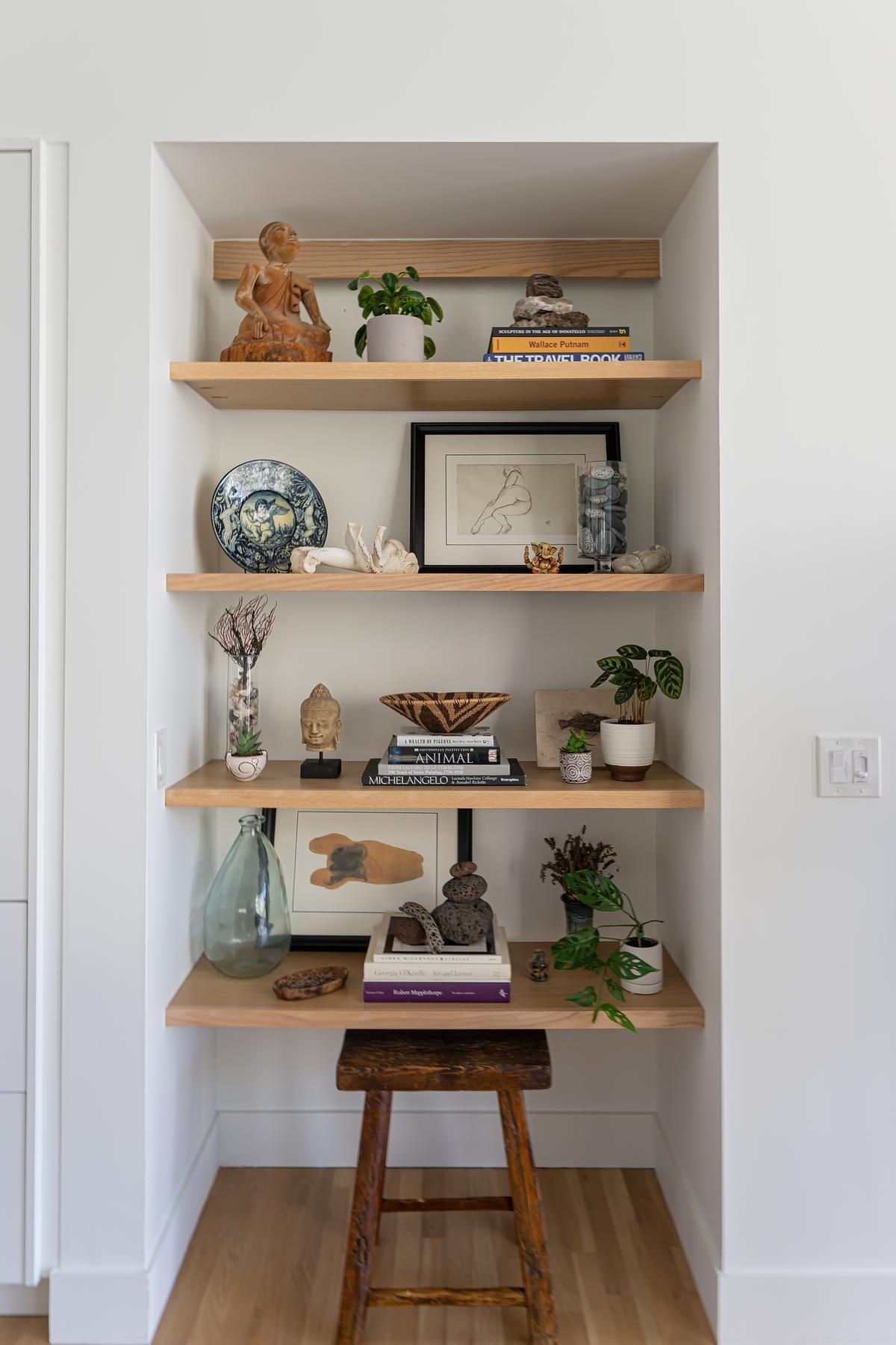 Further into the home, a built-in shelving niche features floating wood shelves. These shelves hold books and decorative items, adding warmth and personality while maintaining the home’s clean, minimal aesthetic.