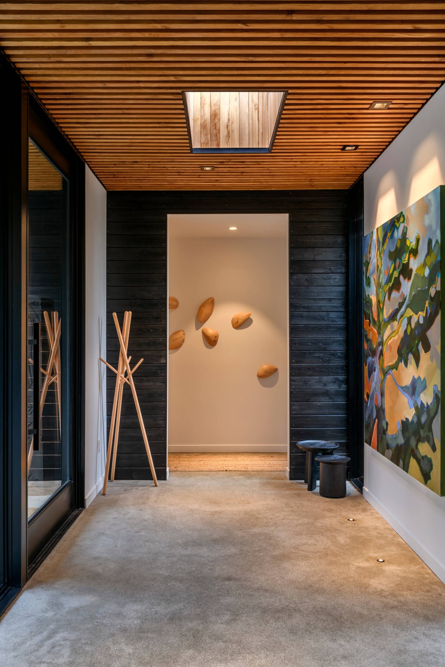 Natural textures and thoughtful details make this hallway feel instantly welcoming. The slatted wood ceiling draws the eye upward, where a skylight softly filters daylight into the space. A mix of matte black and honey-toned wood creates a warm contrast, echoed in the sculptural wall art and simple coat stand.