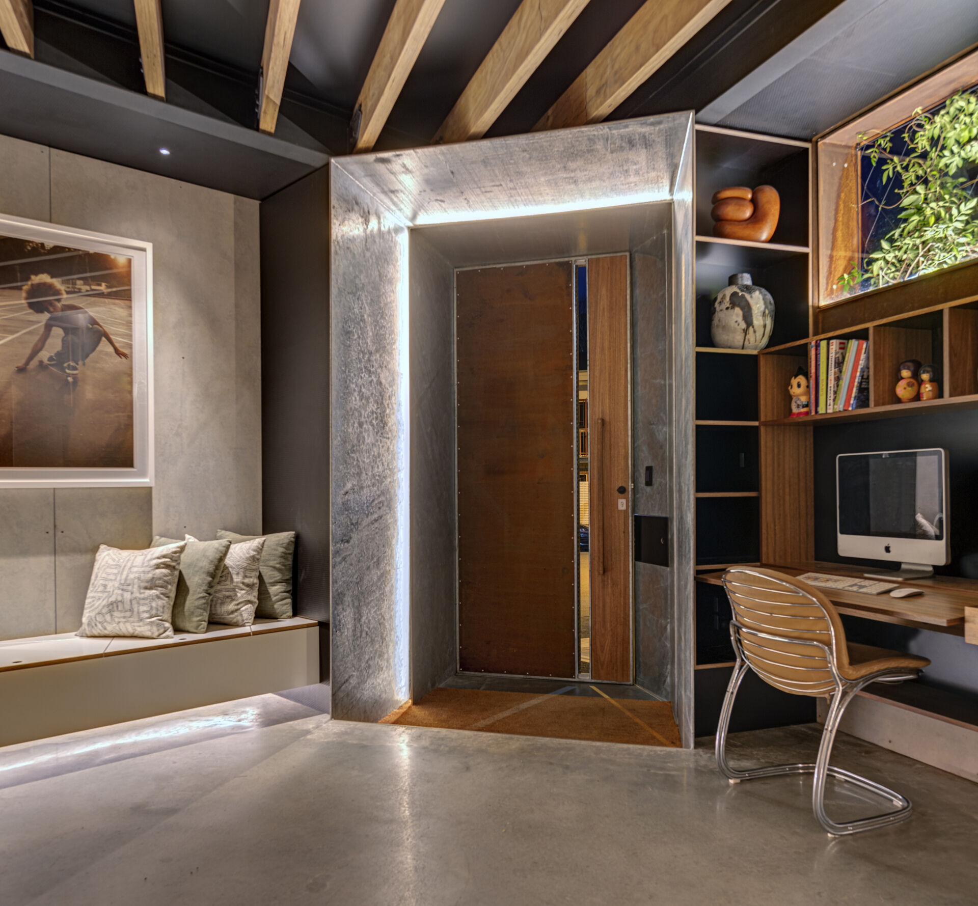 The entryway introduces a layered palette of timber, concrete, and soft accents. A built-in bench with upholstered cushions sits beneath art, while polished concrete floors and rendered walls offer contrast, while exposed ceiling beams reference the building’s industrial past. Soft lighting at the threshold and along the bench adds a low, ambient glow.