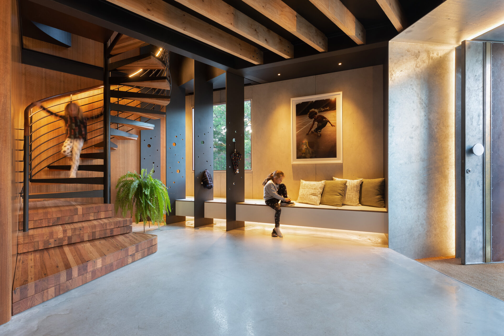 The entryway introduces a layered palette of timber, concrete, and soft accents. A built-in bench with upholstered cushions sits beneath art, while polished concrete floors and rendered walls offer contrast, while exposed ceiling beams reference the building’s industrial past. Soft lighting at the threshold and along the bench adds a low, ambient glow.