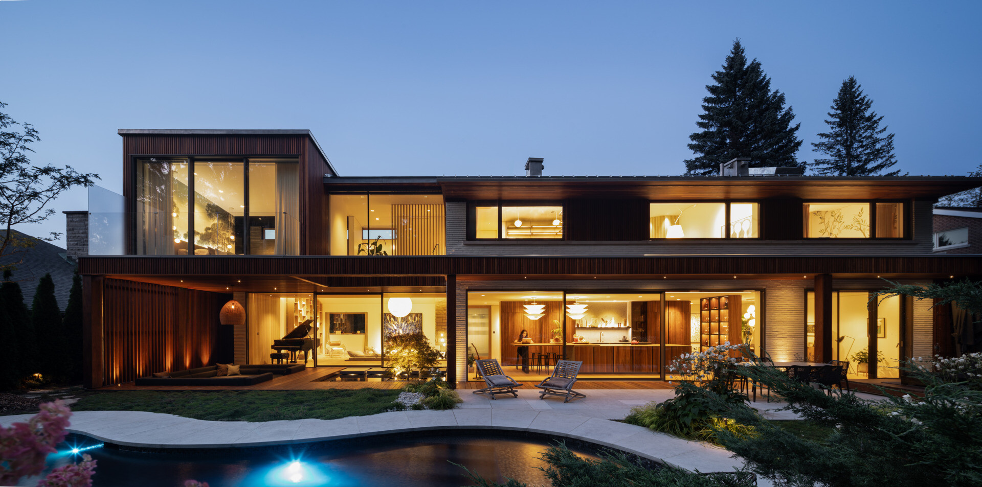 At the back of the home, a sunken outdoor lounge forms a quiet refuge framed by trees. The rear facade, largely glazed, opens up to this landscape, while vertical IPE wood slats echo the forest’s rhythm and provide privacy. Slate roof overhangs shade the space while visually rooting the house into its setting.
