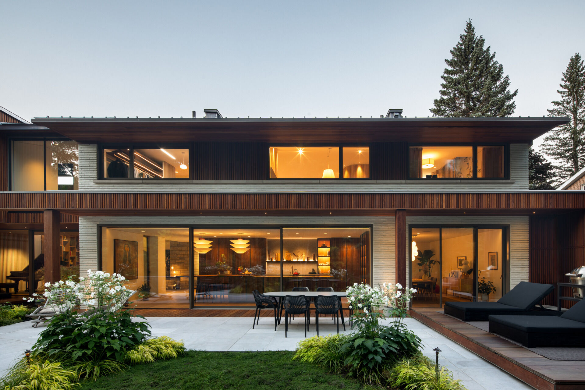 At the back of the home, a sunken outdoor lounge forms a quiet refuge framed by trees. The rear facade, largely glazed, opens up to this landscape, while vertical IPE wood slats echo the forest’s rhythm and provide privacy. Slate roof overhangs shade the space while visually rooting the house into its setting.