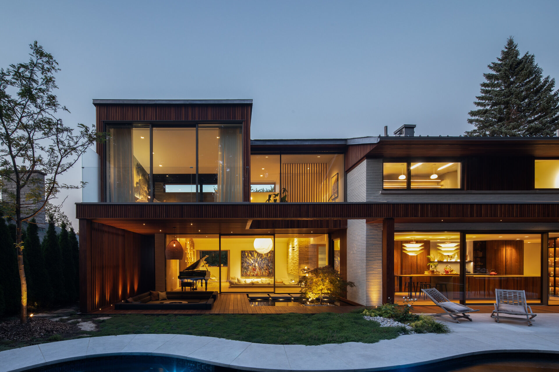 At the back of the home, a sunken outdoor lounge forms a quiet refuge framed by trees. The rear facade, largely glazed, opens up to this landscape, while vertical IPE wood slats echo the forest’s rhythm and provide privacy. Slate roof overhangs shade the space while visually rooting the house into its setting.
