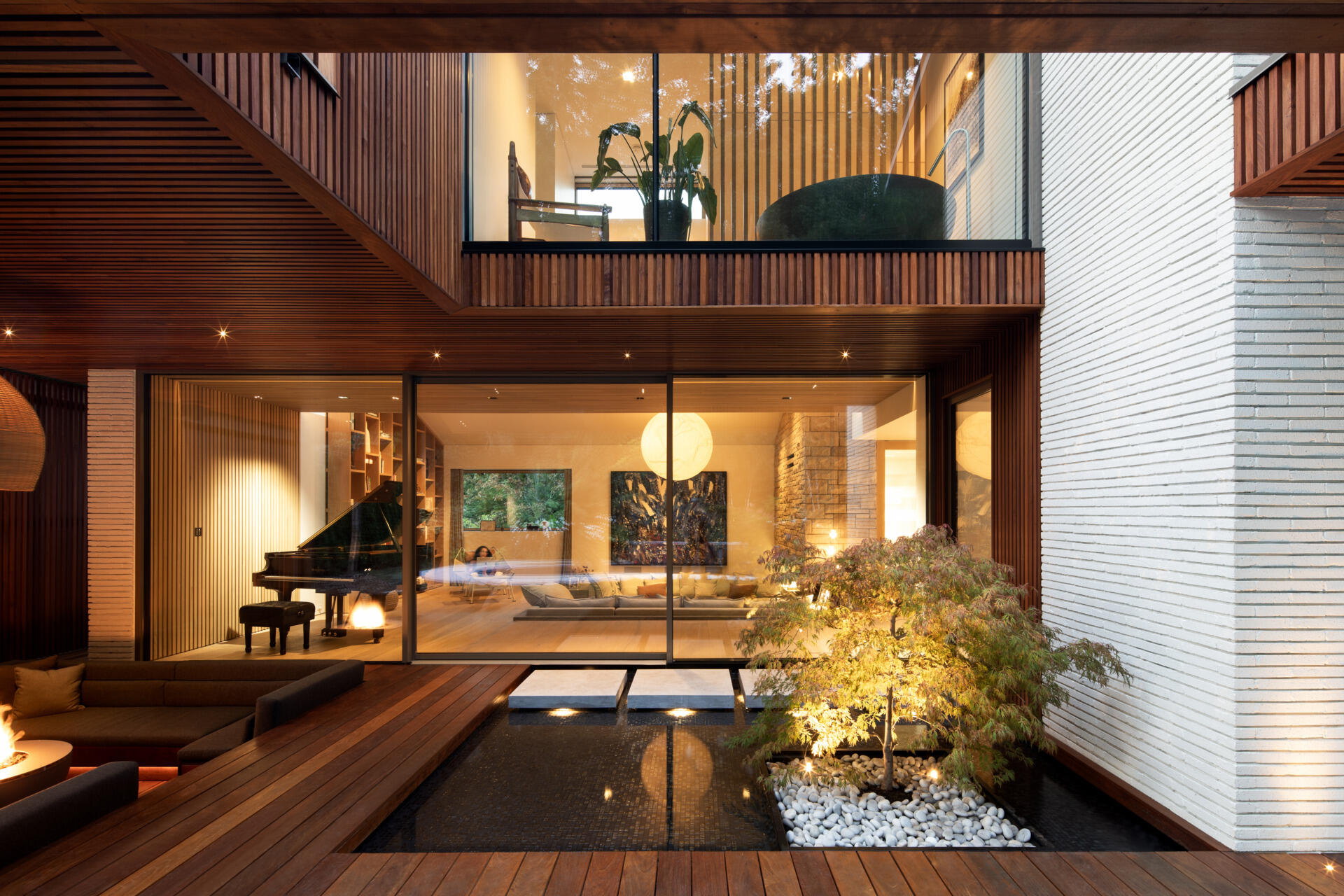 At the back of the home, a sunken outdoor lounge forms a quiet refuge framed by trees. The rear facade, largely glazed, opens up to this landscape, while vertical IPE wood slats echo the forest’s rhythm and provide privacy. Slate roof overhangs shade the space while visually rooting the house into its setting.