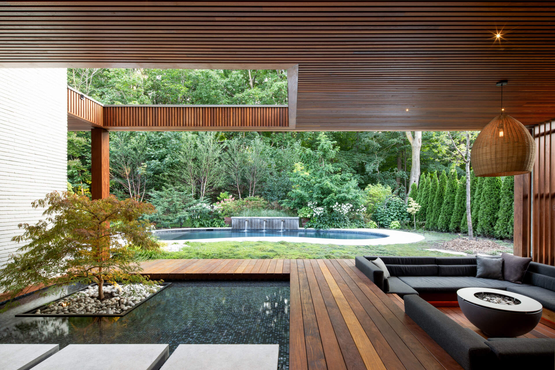 At the back of the home, a sunken outdoor lounge forms a quiet refuge framed by trees. The rear facade, largely glazed, opens up to this landscape, while vertical IPE wood slats echo the forest’s rhythm and provide privacy. Slate roof overhangs shade the space while visually rooting the house into its setting.