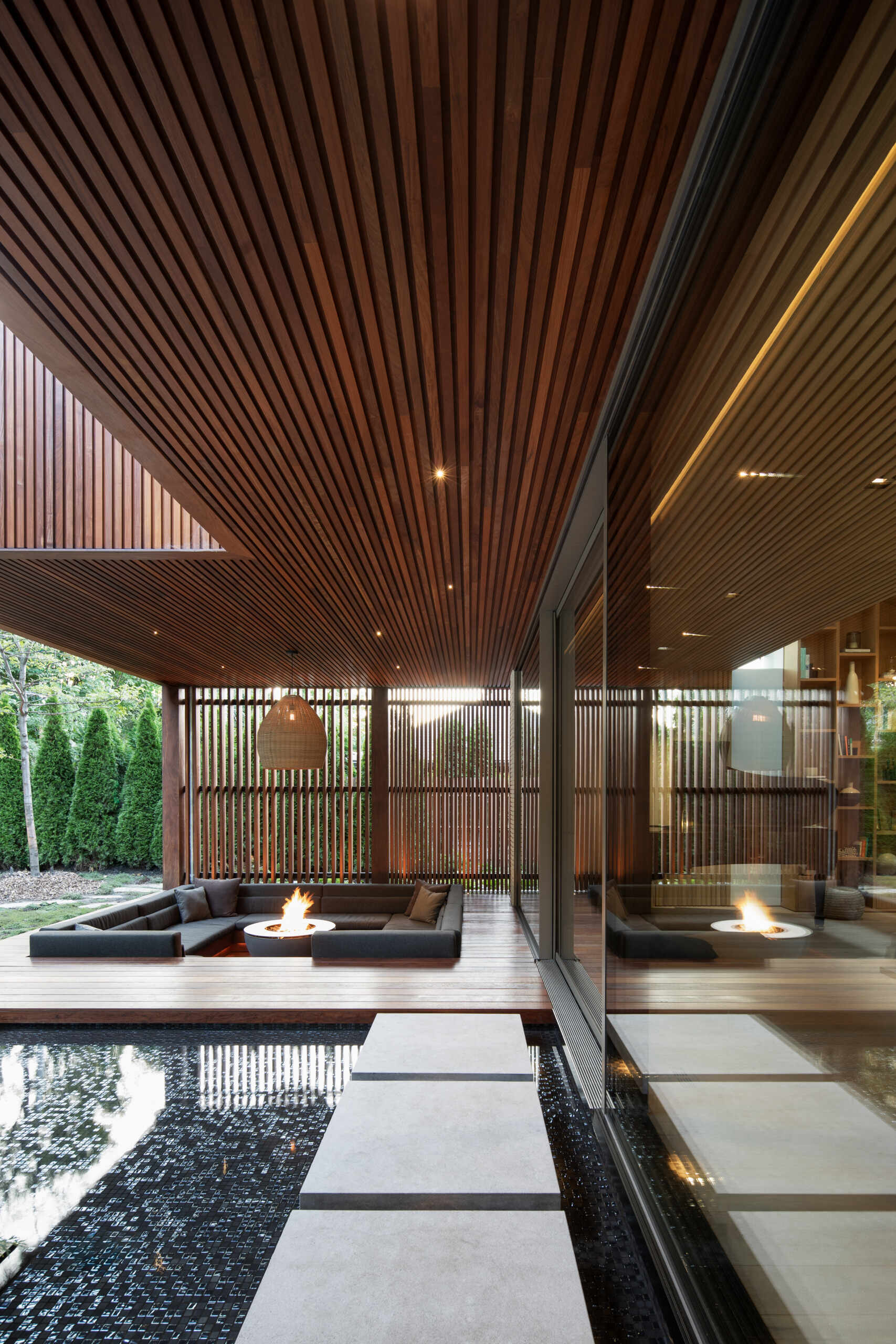 At the back of the home, a sunken outdoor lounge forms a quiet refuge framed by trees. The rear facade, largely glazed, opens up to this landscape, while vertical IPE wood slats echo the forest’s rhythm and provide privacy. Slate roof overhangs shade the space while visually rooting the house into its setting.