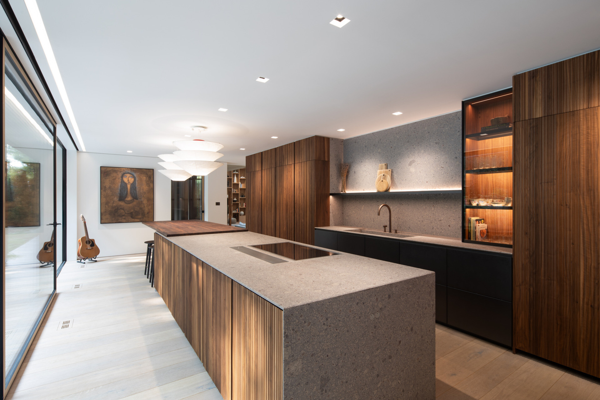 The kitchen blends precision with warmth, anchored by a sculptural island in textured stone and fluted walnut. A muted palette of grey, black, and bronze gives the space a grounded elegance. Integrated lighting highlights a recessed shelf and a glass-fronted pantry, while the oversized pendants above the dining bar adds softness and scale.