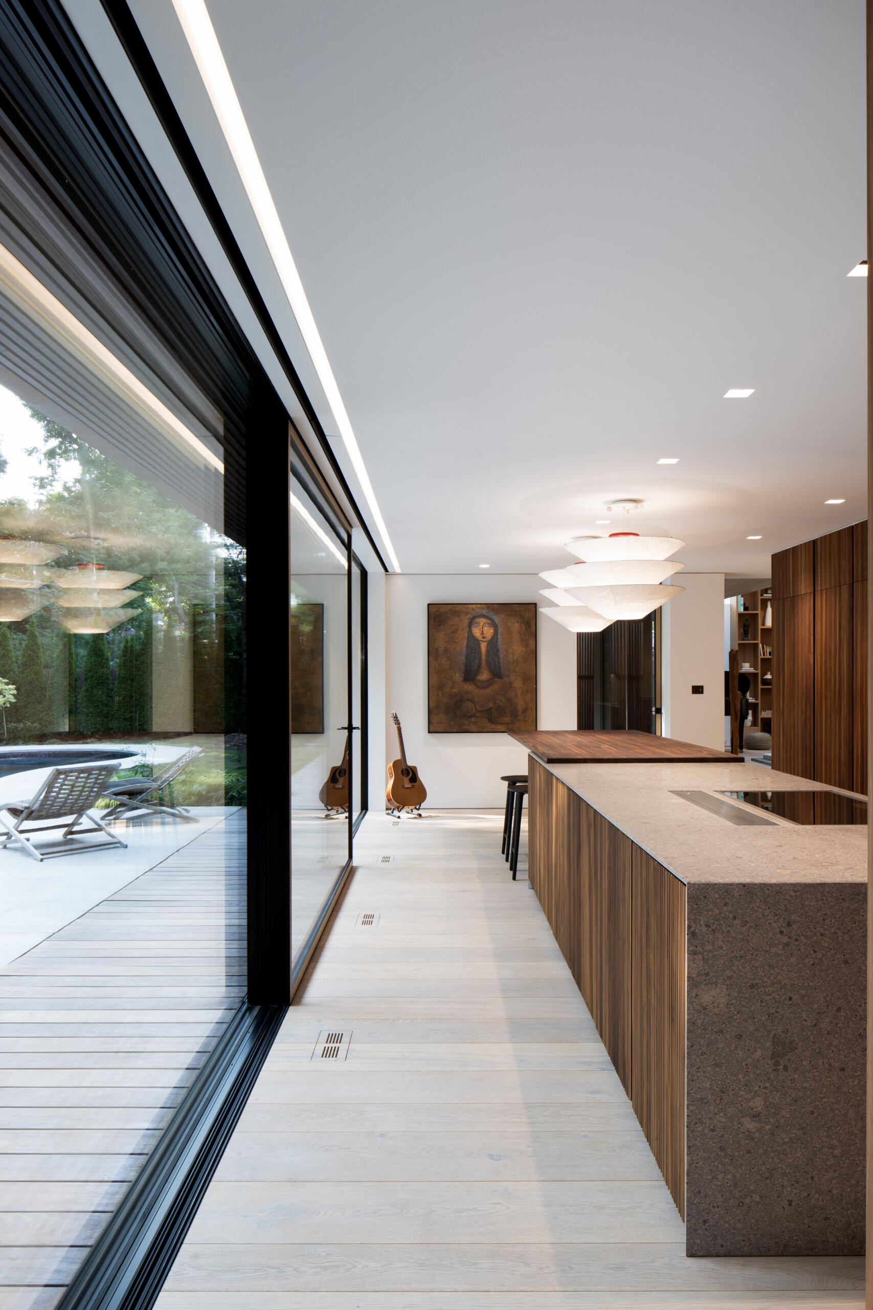 The kitchen blends precision with warmth, anchored by a sculptural island in textured stone and fluted walnut. A muted palette of grey, black, and bronze gives the space a grounded elegance. Integrated lighting highlights a recessed shelf and a glass-fronted pantry, while the oversized pendants above the dining bar adds softness and scale.