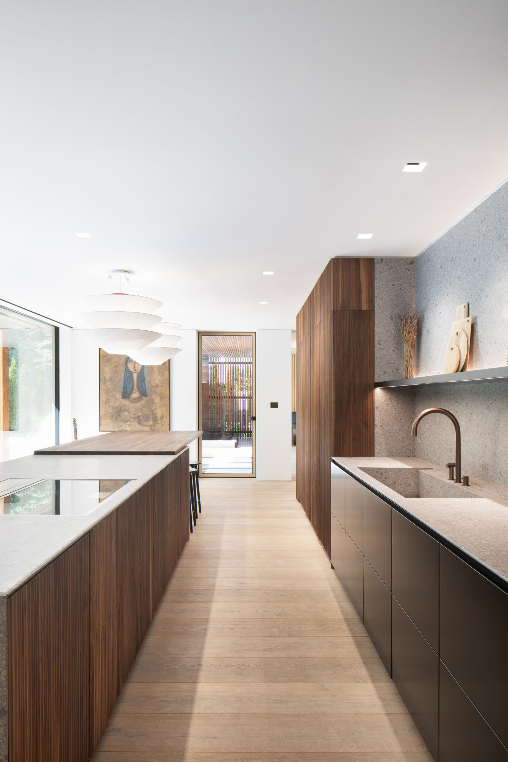 The kitchen blends precision with warmth, anchored by a sculptural island in textured stone and fluted walnut. A muted palette of grey, black, and bronze gives the space a grounded elegance. Integrated lighting highlights a recessed shelf and a glass-fronted pantry, while the oversized pendants above the dining bar adds softness and scale.