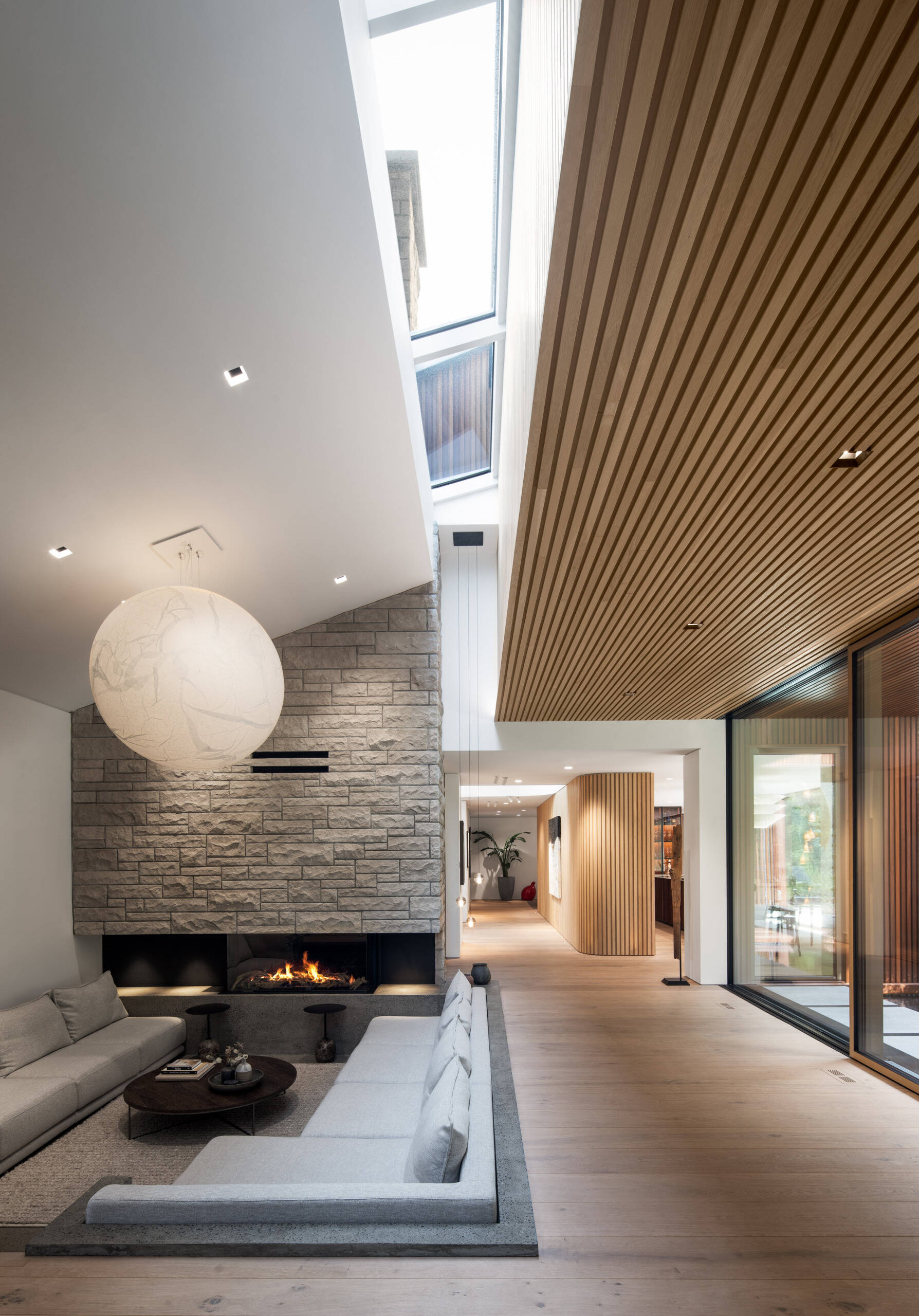 Inside, the living area is a lesson in restraint. Natural oak and walnut meet stone and marble, with no baseboards or moldings in sight. The double-height cathedral ceiling brings a sense of drama, softened by curves and textured surfaces. Hidden doors and architectural lighting preserve the visual flow, letting materiality and form do the talking.