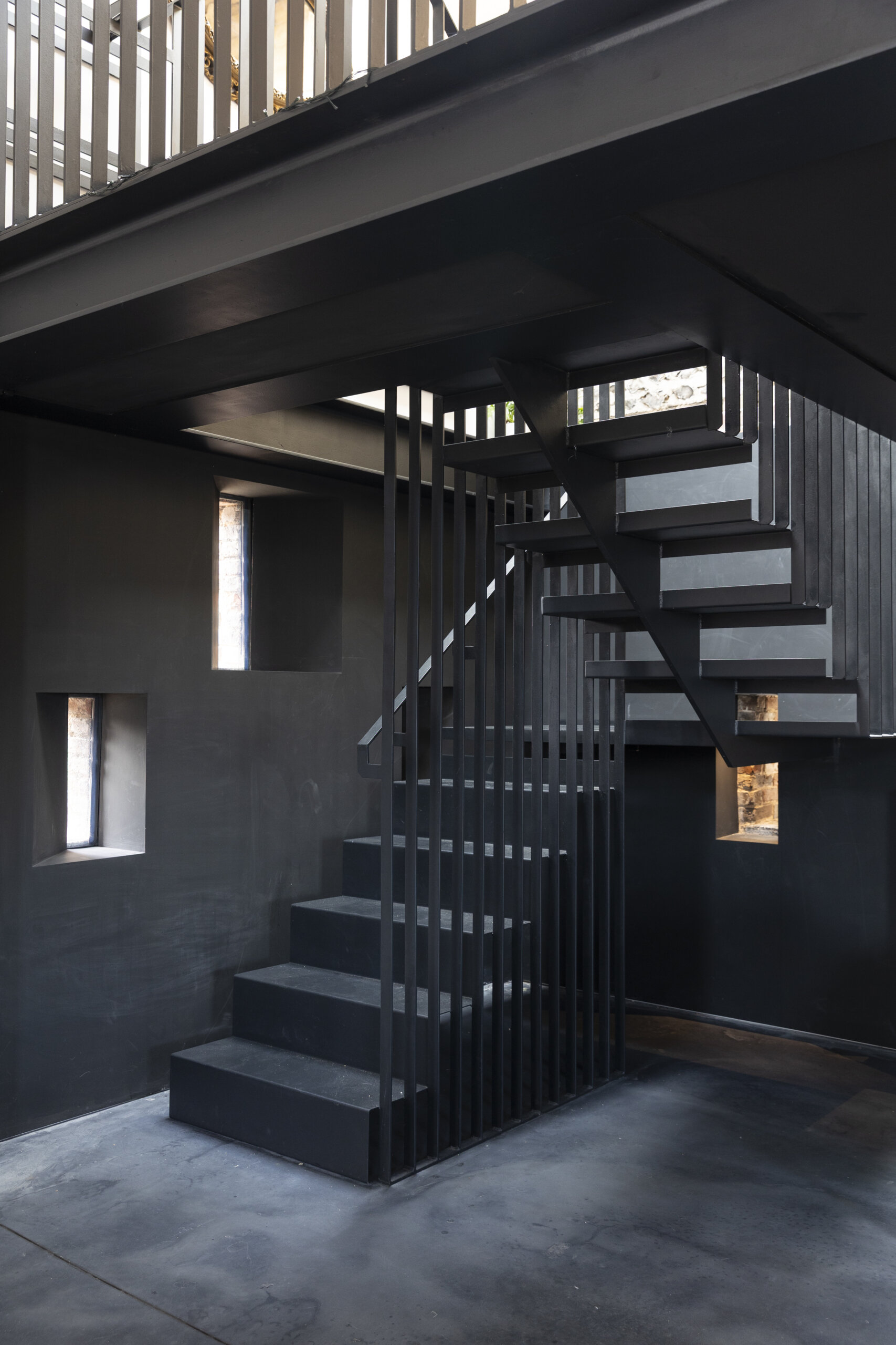 The bespoke steel staircase doubles as a sculptural centerpiece and a functional link between levels. Nearby, the library tucks into a quieter corner of the home, framed by matte black walls and flooded with soft natural light from small windows.