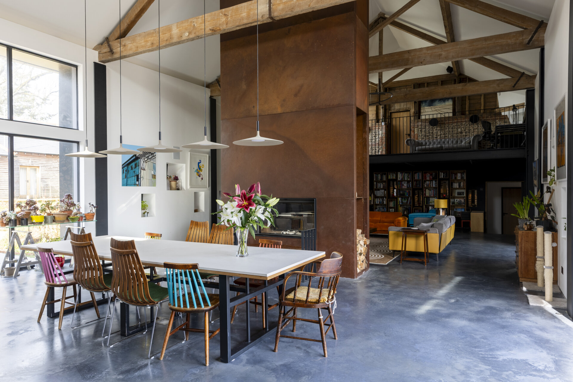 At the center of the home, the living room is anchored by a corten steel fireplace, an architectural statement that radiates both heat and visual weight. Above, a mezzanine level floats within the double-height space, framed by original timber beams.