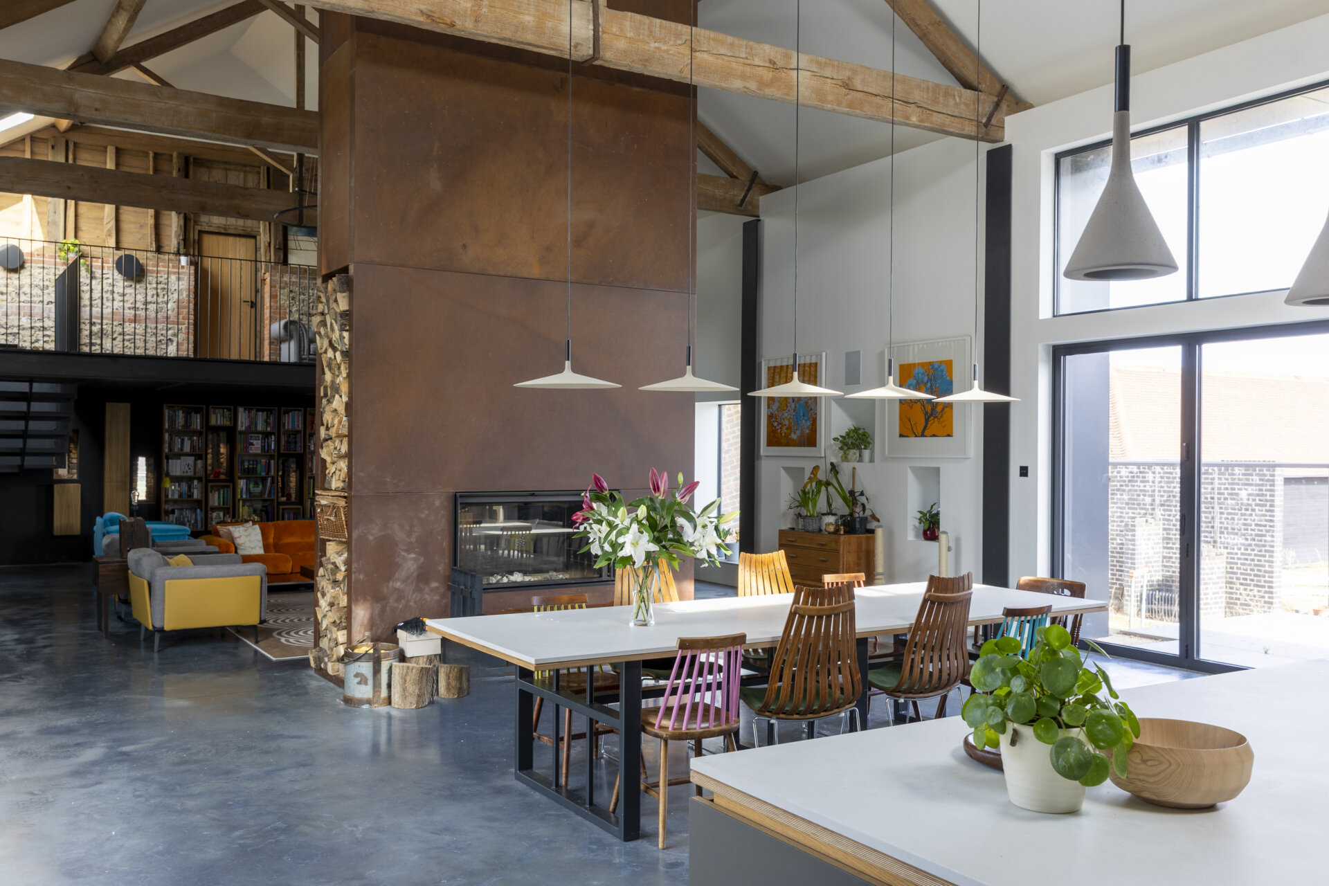 At the center of the home, the living room is anchored by a corten steel fireplace, an architectural statement that radiates both heat and visual weight. Above, a mezzanine level floats within the double-height space, framed by original timber beams.