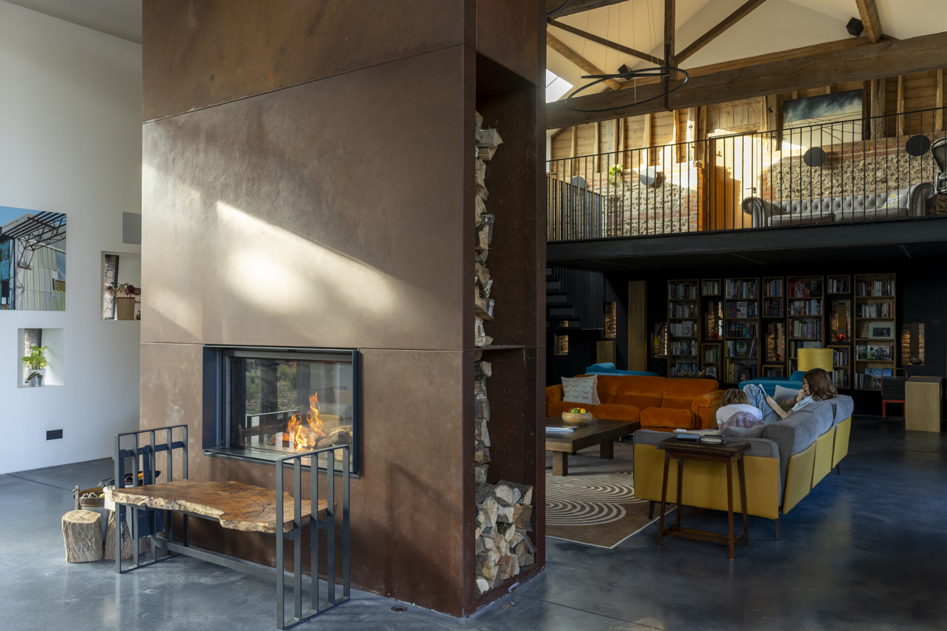 At the center of the home, the living room is anchored by a corten steel fireplace, an architectural statement that radiates both heat and visual weight. Above, a mezzanine level floats within the double-height space, framed by original timber beams.