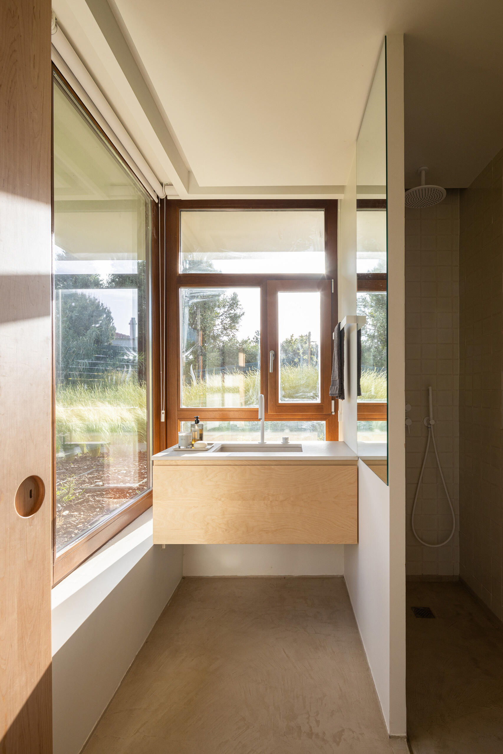 The bathroom embraces the tactile simplicity that defines the rest of the home. Soft light and natural tones define the bathroom, where creamy walls, pale wood, and smooth concrete create a grounded calm. A large window draws the outdoors in, making space for quiet, unhurried mornings.