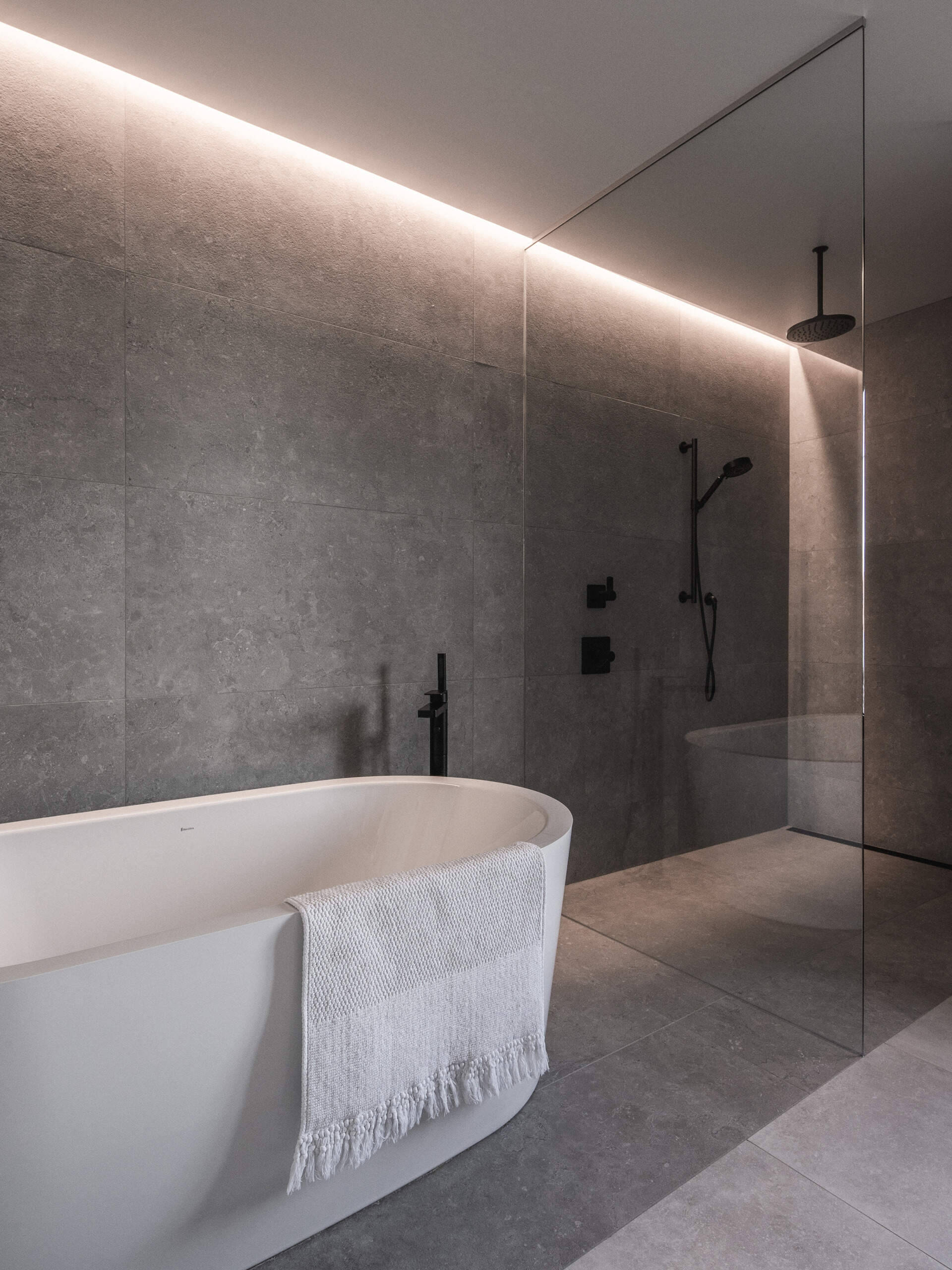 In this bathroom there's a serene and luxurious atmosphere. The space features a freestanding matte white bathtub, while the walls and floors are clad in large-format gray stone tiles that create a cohesive, monolithic look. A frameless glass partition separates the wet area, which includes a walk-in shower with a ceiling-mounted rainfall showerhead and a matte black hand shower set.
