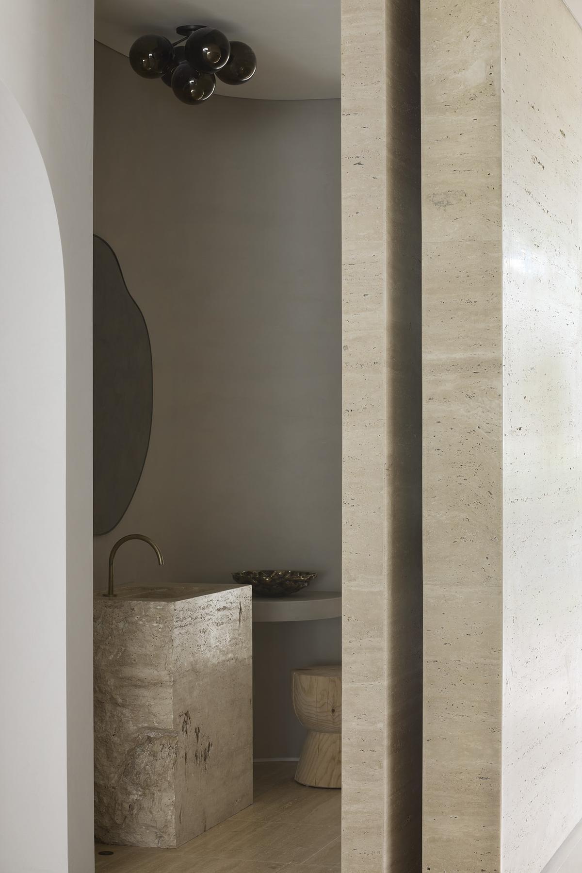 This powder room pairs striking materials with sculptural minimalism. A raw stone pedestal sink grounds the space with its natural texture, while soft, curved walls and warm gray tones create a calming cocoon. The smoky glass light fixture overhead adds a moody glow, and the irregular mirror shape brings a touch of whimsy.