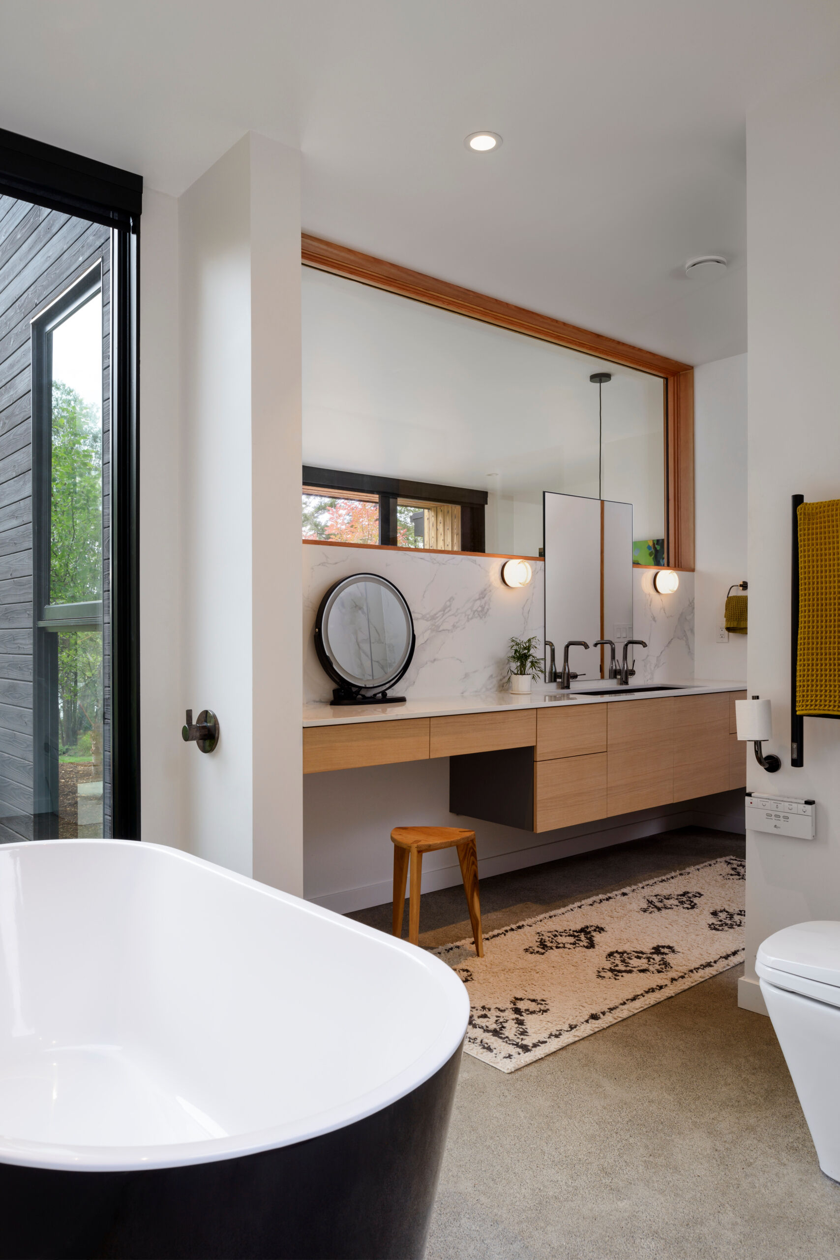 This modern bathroom blends clean lines with natural textures, featuring a floating wood vanity, soaking tub, and views into the surrounding trees.