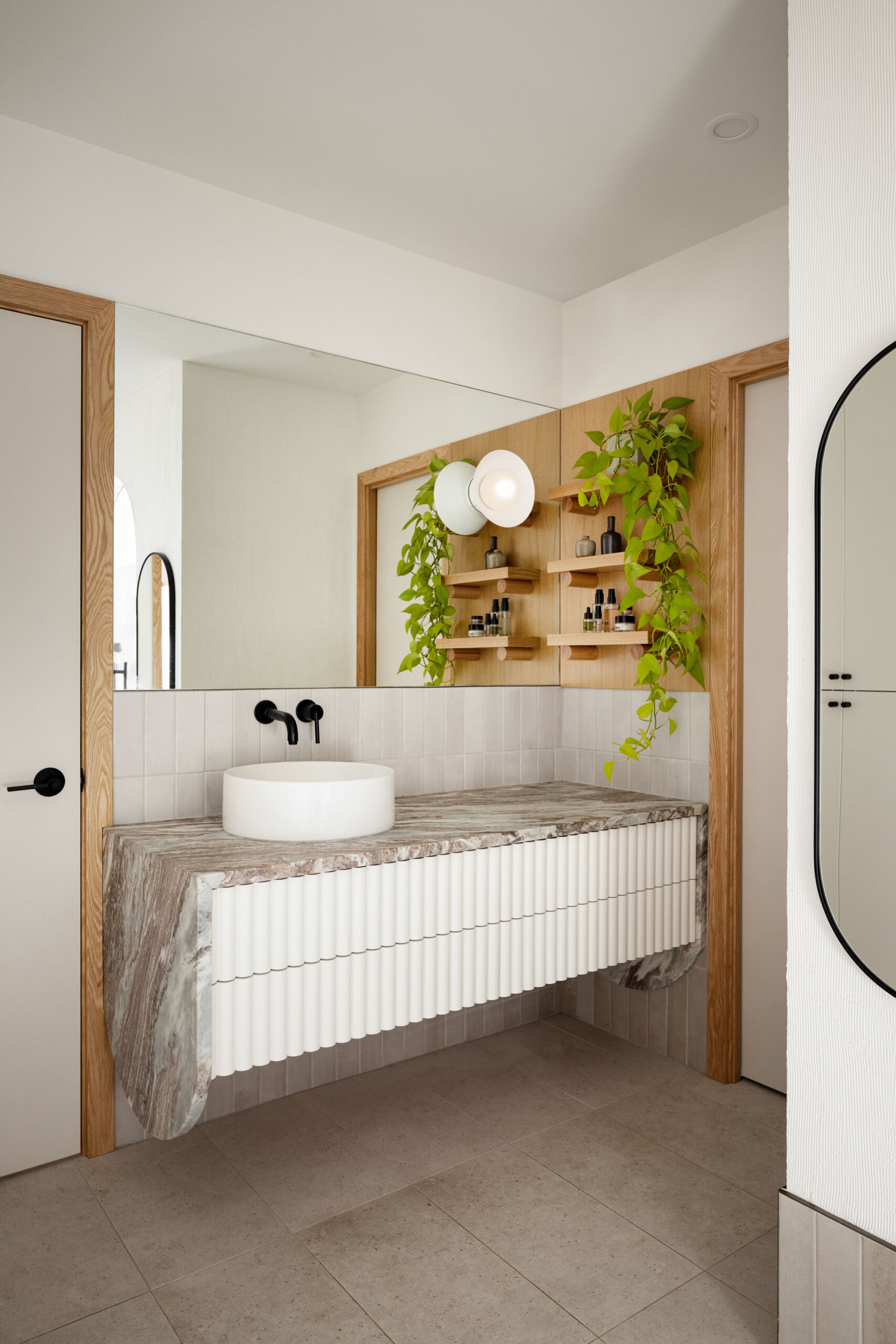 This primary bathroom feels beautifully calm with its creamy tile walls and veined stone countertop, anchored by a fluted white vanity that adds texture and interest. Matte black fixtures give a modern contrast, while gentle light from the sconce makes the space feel welcoming.