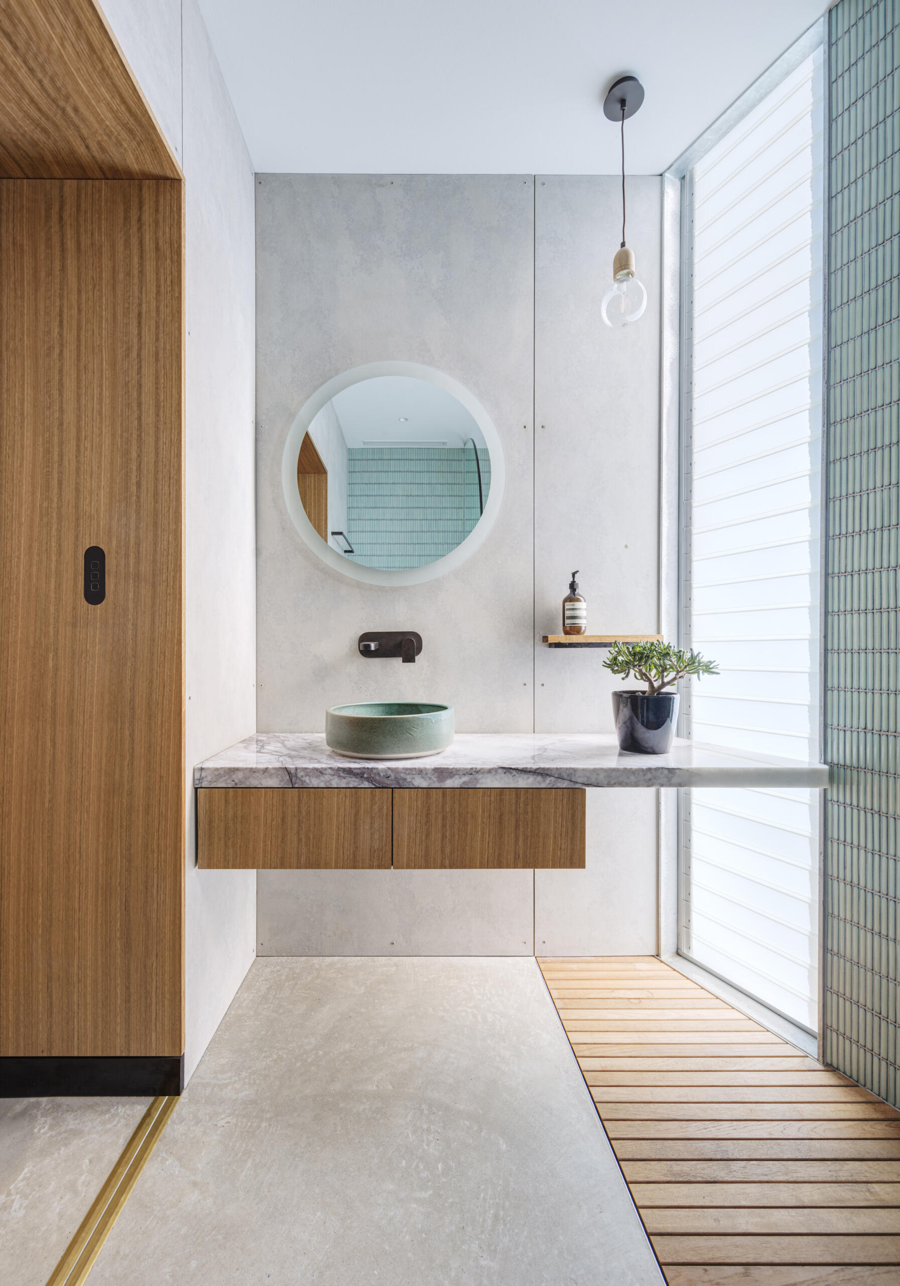 This bathroom is shaped by light, texture, and restraint. Louvered windows allows soft daylight to filter in, casting subtle patterns across smooth concrete walls. A floating marble vanity introduces a sense of lightness, grounded by warm timber drawers and flooring. A pale green vessel basin offers a quiet accent, complemented by a round mirror and a single potted plant.
