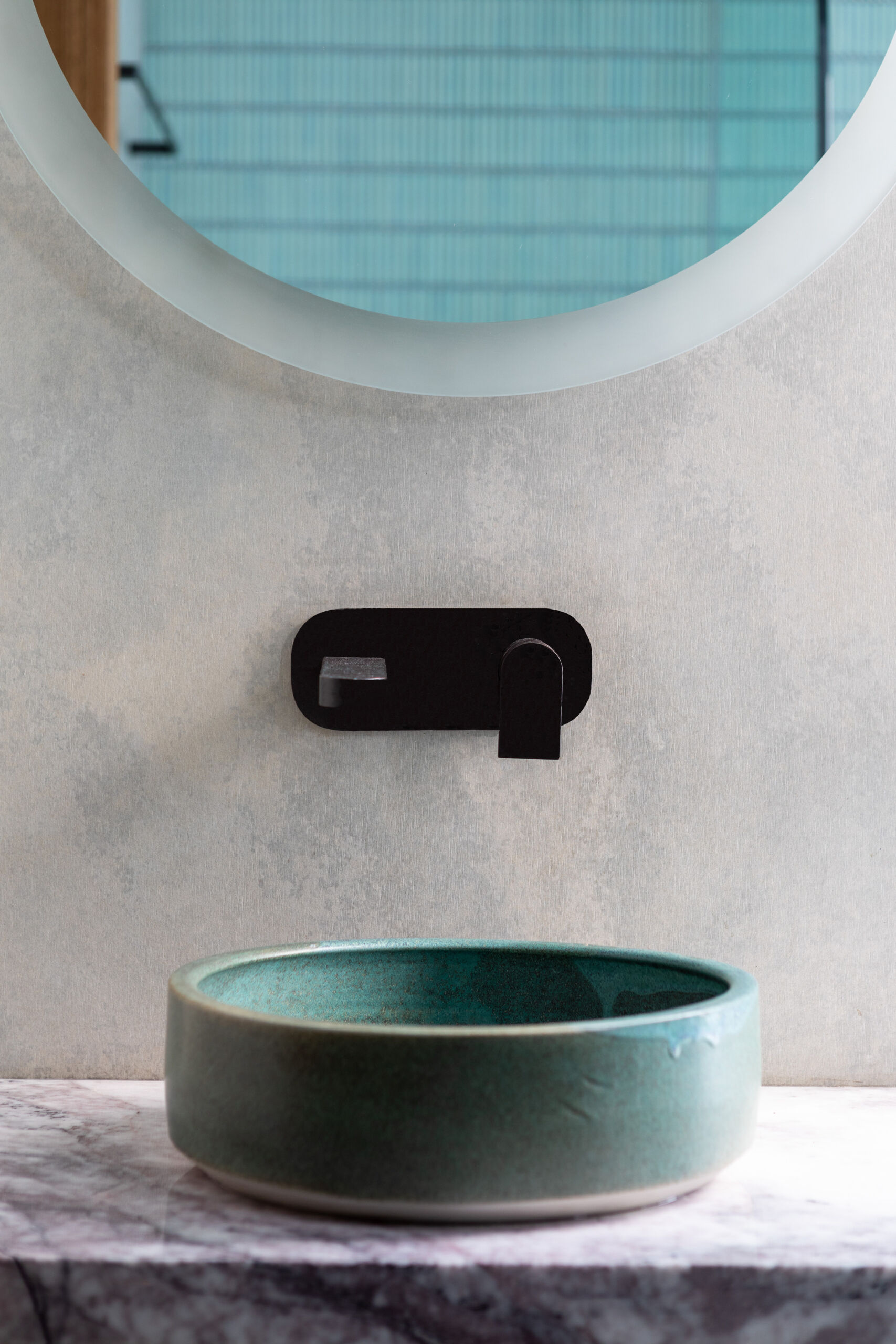This bathroom is shaped by light, texture, and restraint. Louvered windows allows soft daylight to filter in, casting subtle patterns across smooth concrete walls. A floating marble vanity introduces a sense of lightness, grounded by warm timber drawers and flooring. A pale green vessel basin offers a quiet accent, complemented by a round mirror and a single potted plant.