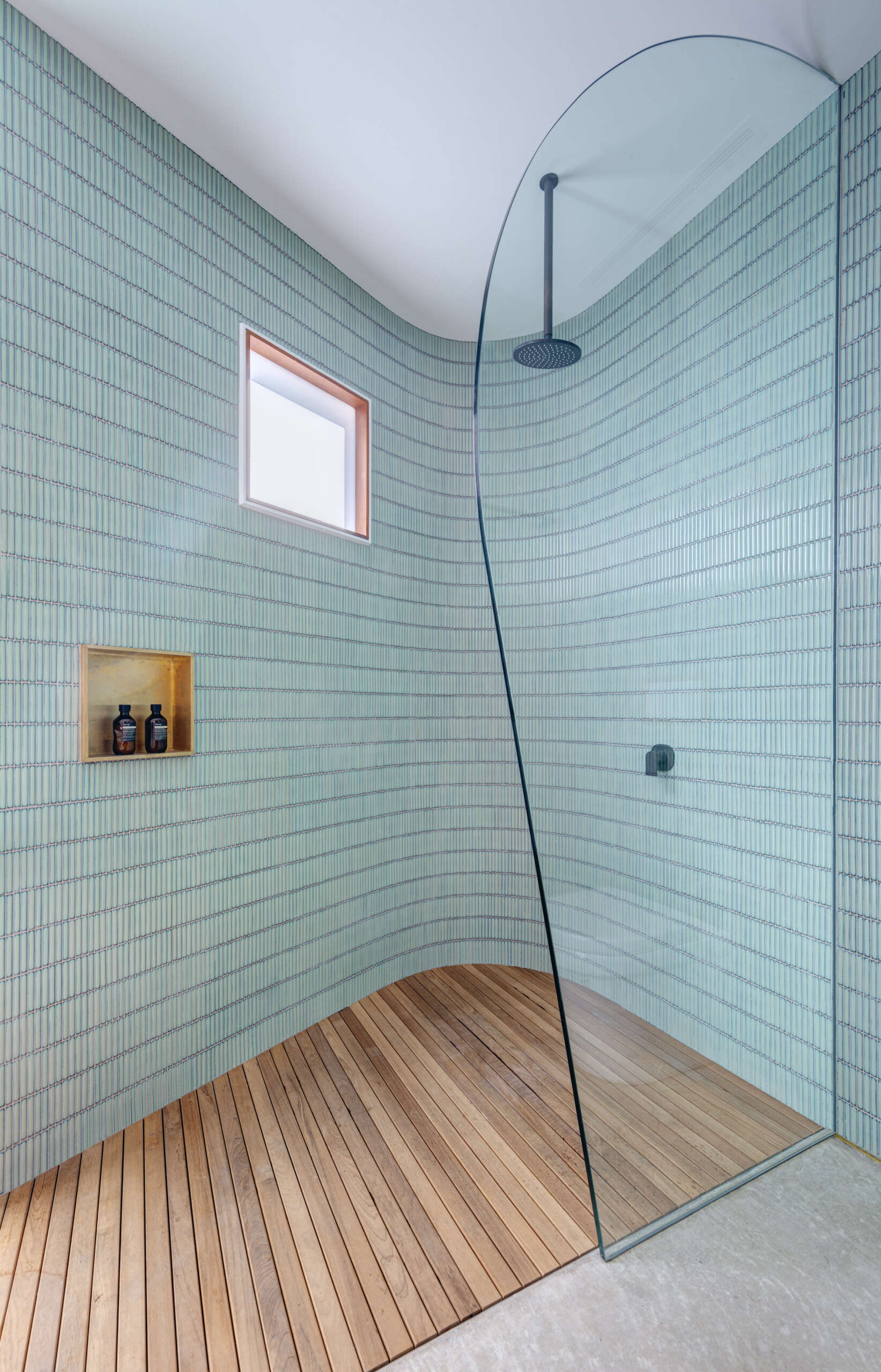 The shower continues the calm material palette. Blue-green vertical tiles wrap gently around the walls, paired with a curved glass screen and streamlined rainfall fixture. A timber slatted floor brings warmth underfoot, while a simple window invites in natural light, reinforcing the room’s quiet, retreat-like quality.