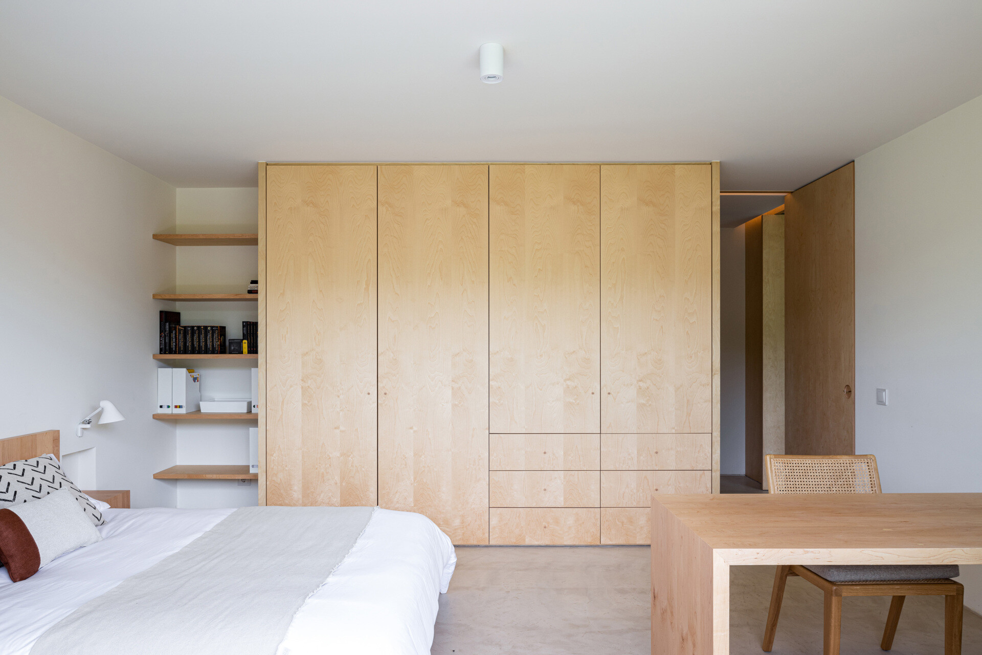 The bedroom is a continuation of calm with wood accents like built-in bedside tables, open shelving, and a desk, while large windows draw nature in.