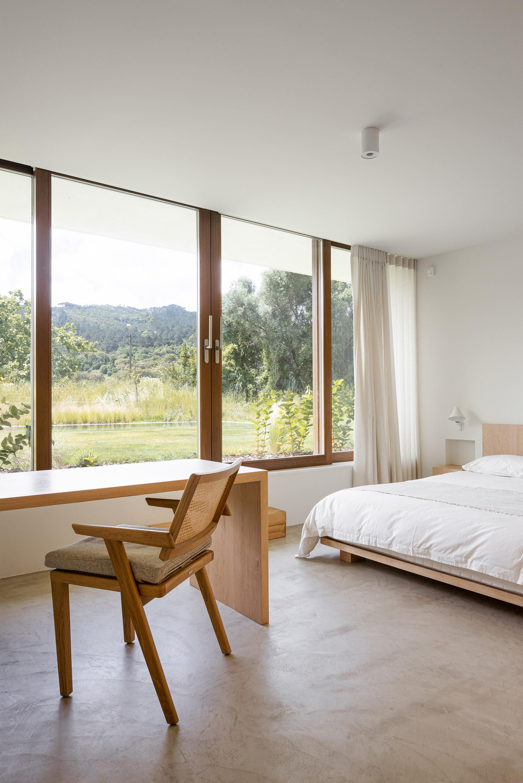 The bedroom is a continuation of calm with wood accents like built-in bedside tables, open shelving, and a desk, while large windows draw nature in.