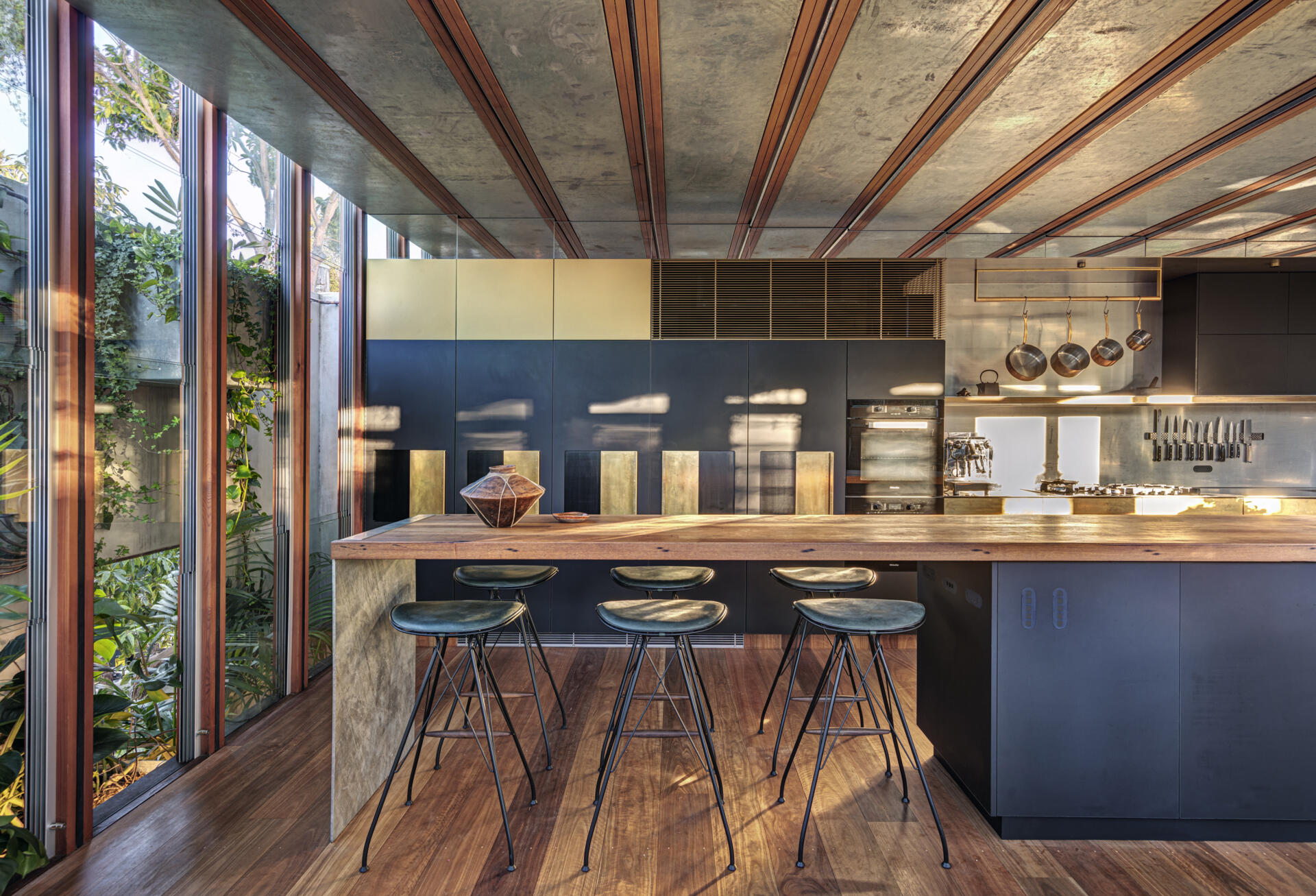 The kitchen pairs dark cabinetry with a timber island, creating a simple, grounded composition. Tall, narrow windows bring in natural light and views of surrounding greenery. Open shelving and brass details add utility without clutter, balancing function and warmth in a space designed for both everyday use and shared moments.