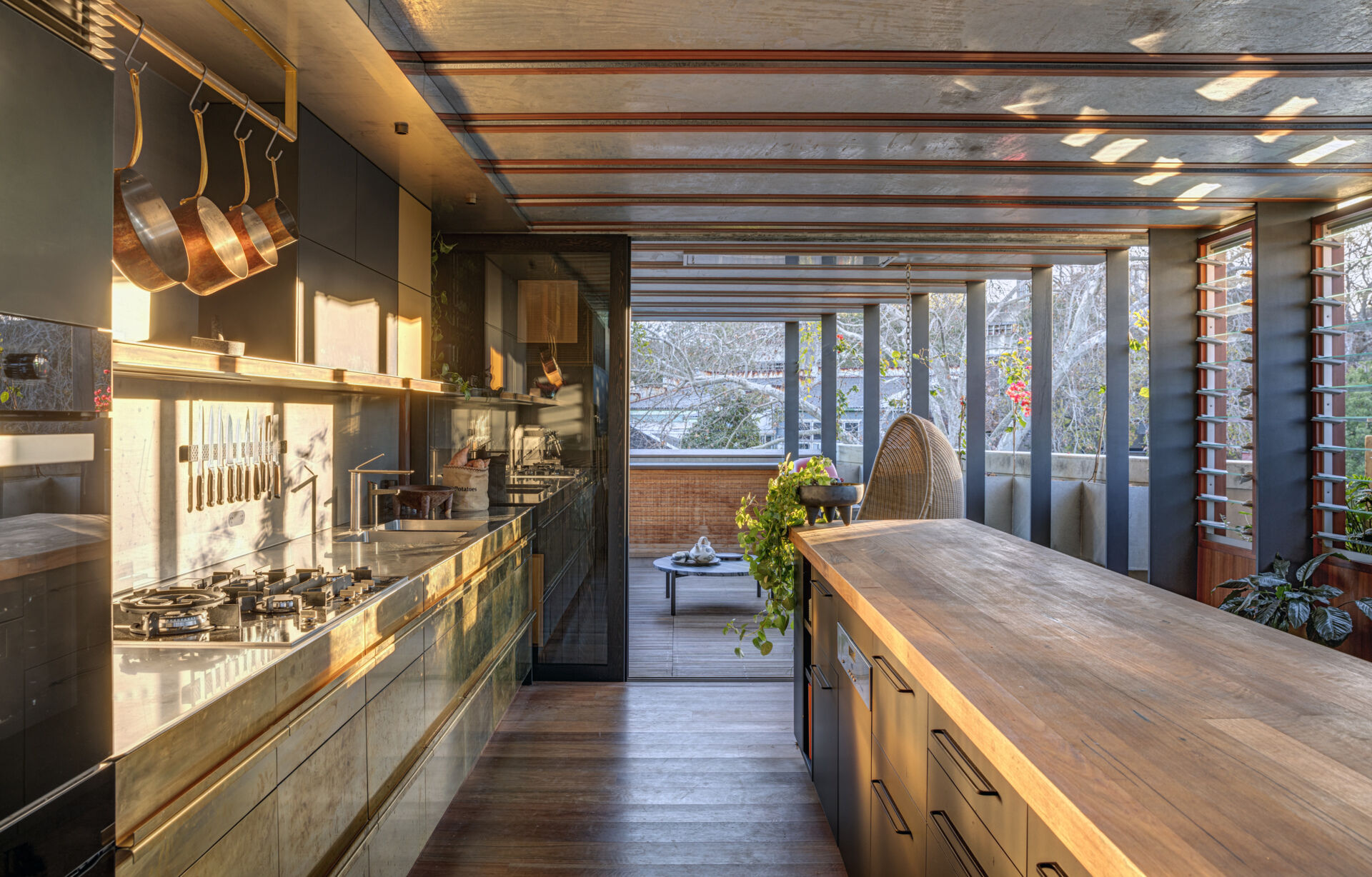 The kitchen pairs dark cabinetry with a timber island, creating a simple, grounded composition. Tall, narrow windows bring in natural light and views of surrounding greenery. Open shelving and brass details add utility without clutter, balancing function and warmth in a space designed for both everyday use and shared moments.